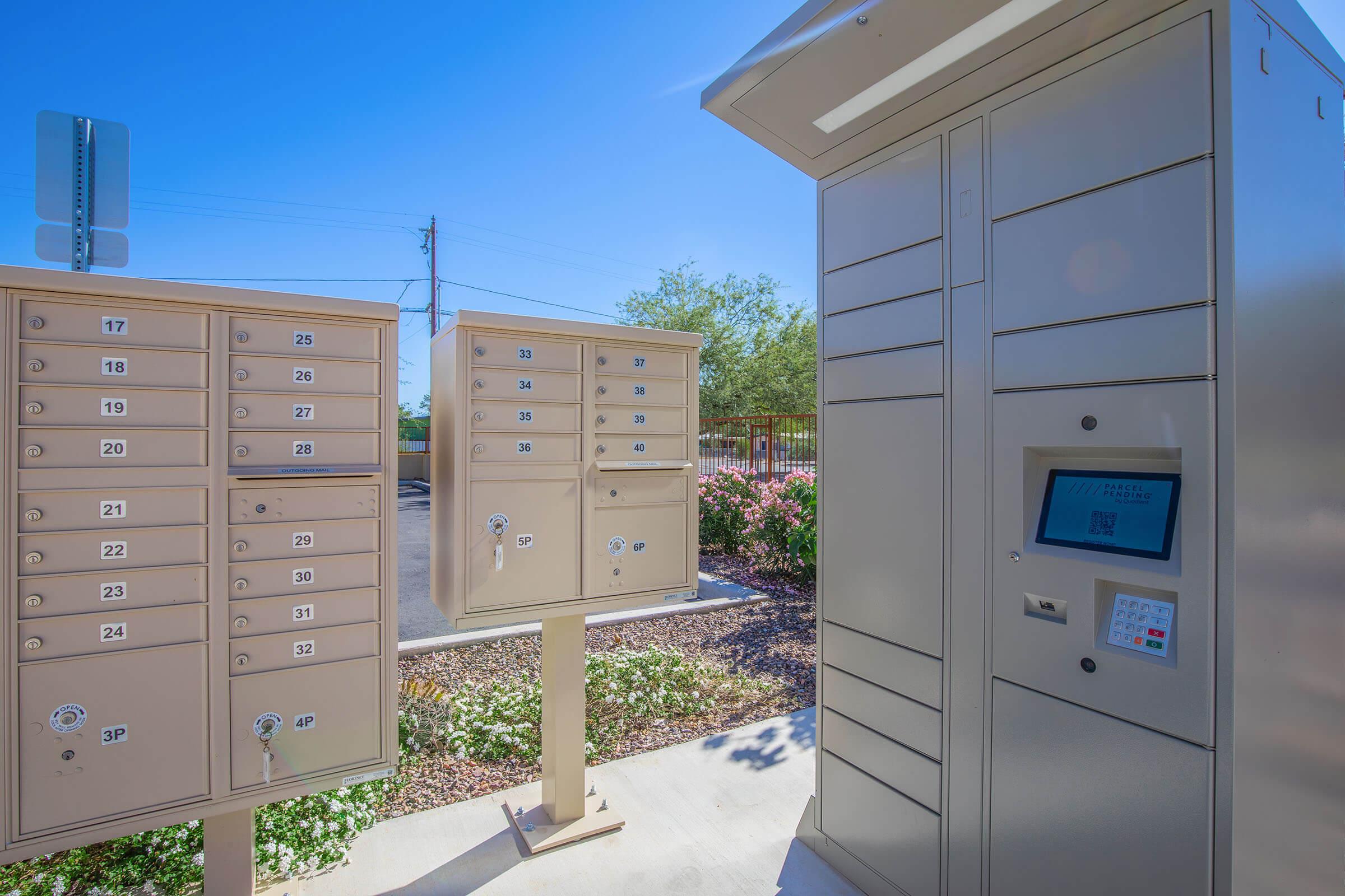 Two clusters of mailboxes in a community setting. On the left, a vertical bank of smaller boxes with numbers ranging from 17 to 36. On the right, a taller mailbox unit with a digital screen and keypad. Surrounding greenery and a clear blue sky enhance the outdoor environment.