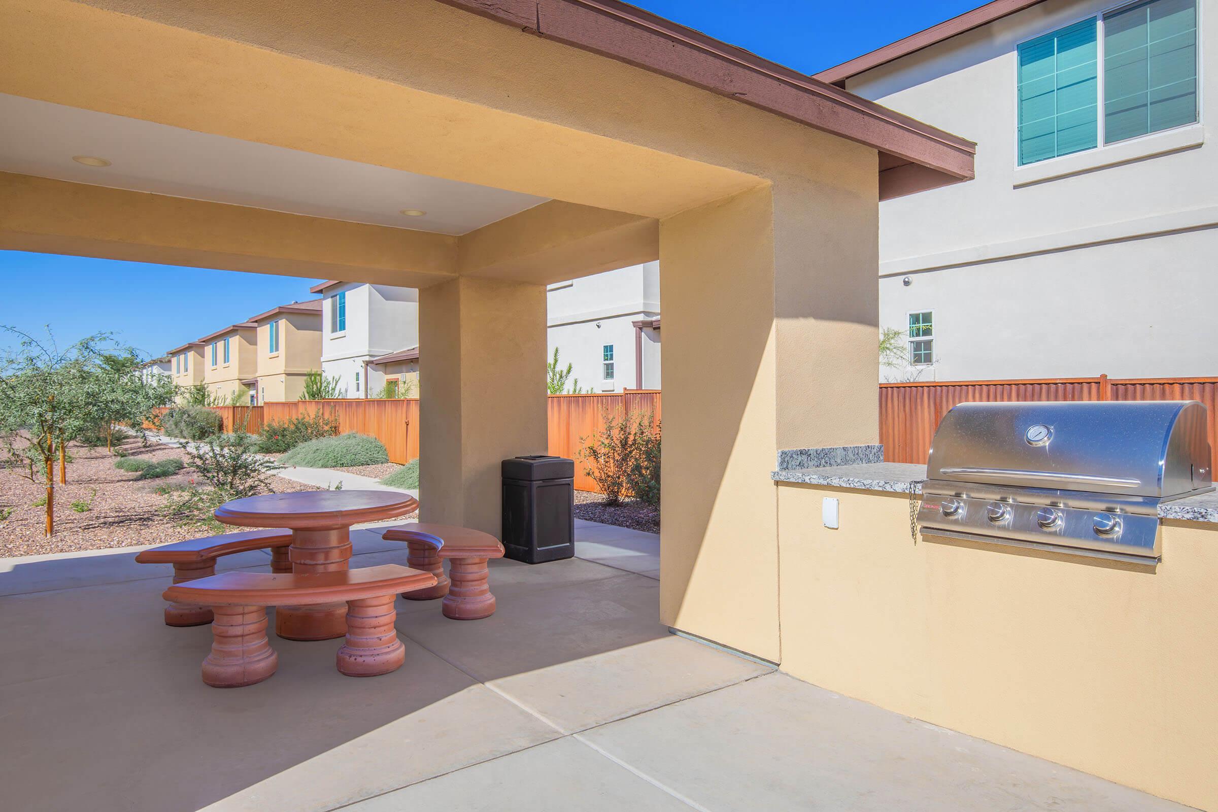 Outdoor patio area featuring a circular picnic table made of stone, a stainless steel grill, and a trash can. The background includes landscaped desert vegetation and residential buildings. The space is bright and inviting, perfect for gatherings and outdoor cooking. Clear blue sky overhead.