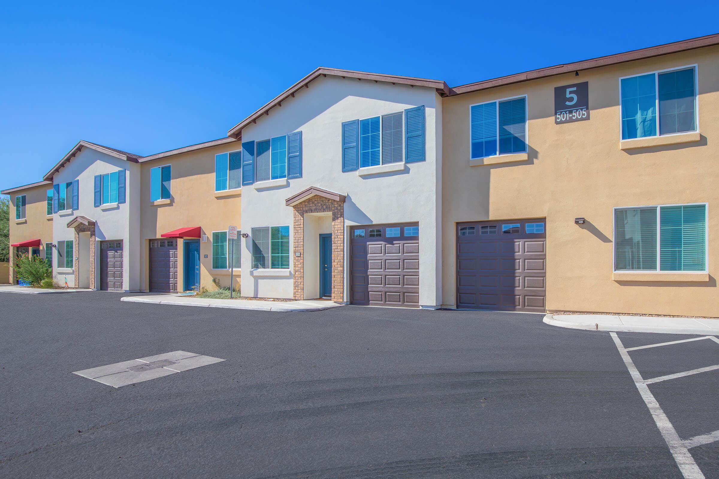 Row of modern townhouses with beige and cream-colored exteriors, blue window shutters, and red awnings. The garages are located at the ground level, and they have a clean, paved parking area. The sky is clear and bright blue, indicating a sunny day.