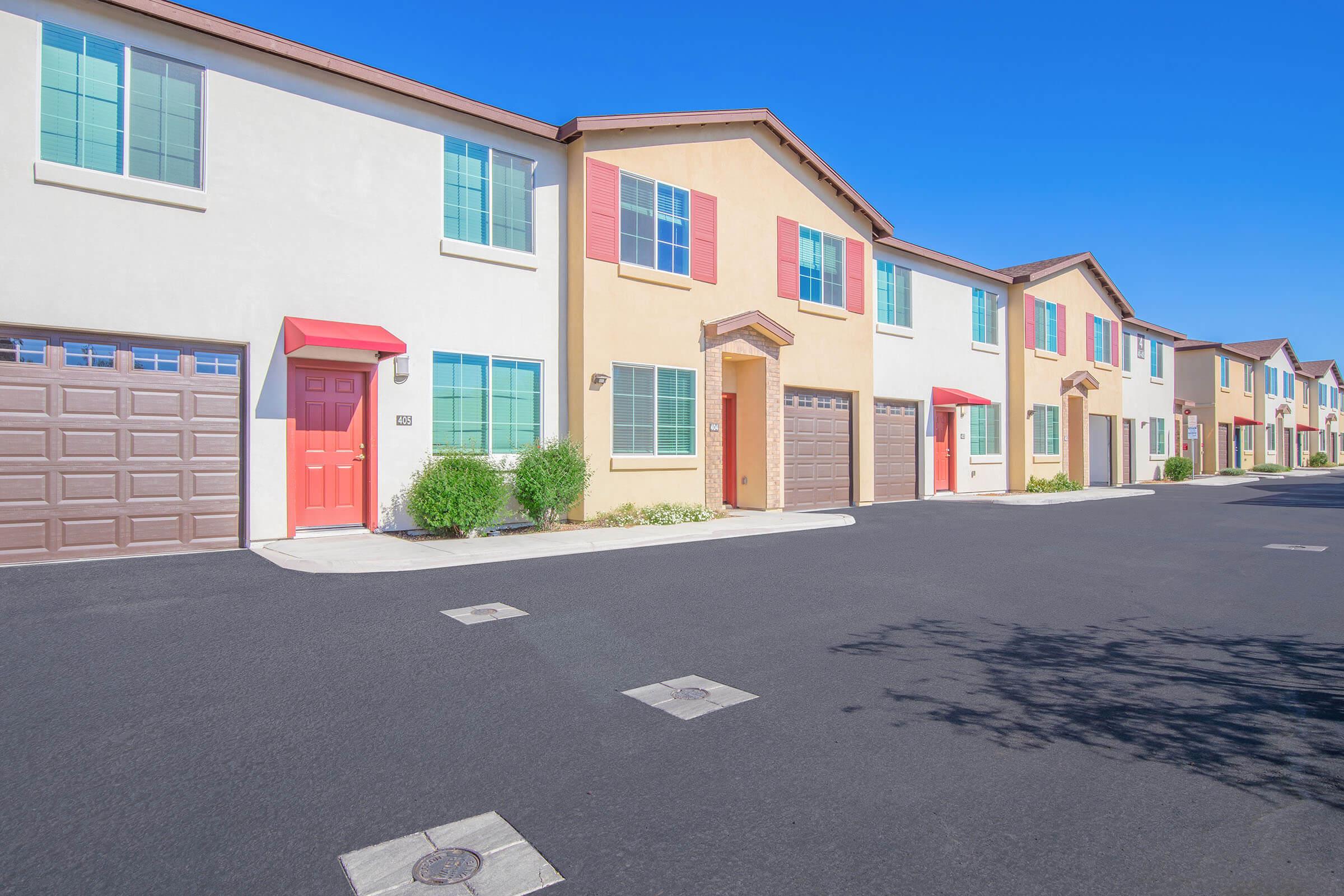 Colorful row of modern townhouses featuring beige and light yellow exteriors, each with distinct front doors and garage doors. The street is clean and well-maintained, bordered by small shrubs under a clear blue sky.