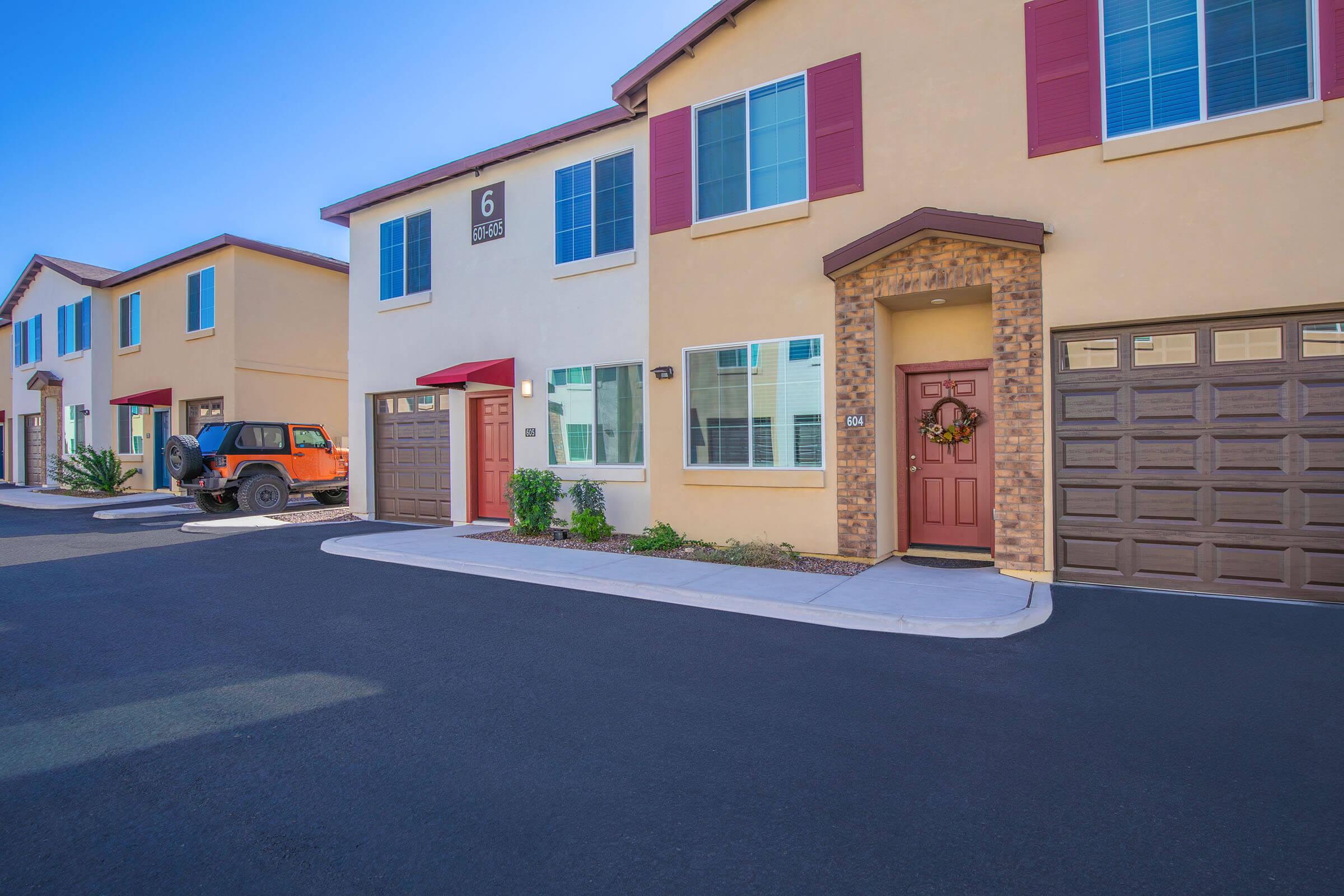 A row of modern, two-story townhouses with beige exteriors and red accents. The first unit features a red door adorned with a wreath, while the garage door is brown. A black SUV is parked in front of one unit. The driveway is paved, and the sky is clear with bright sunlight.