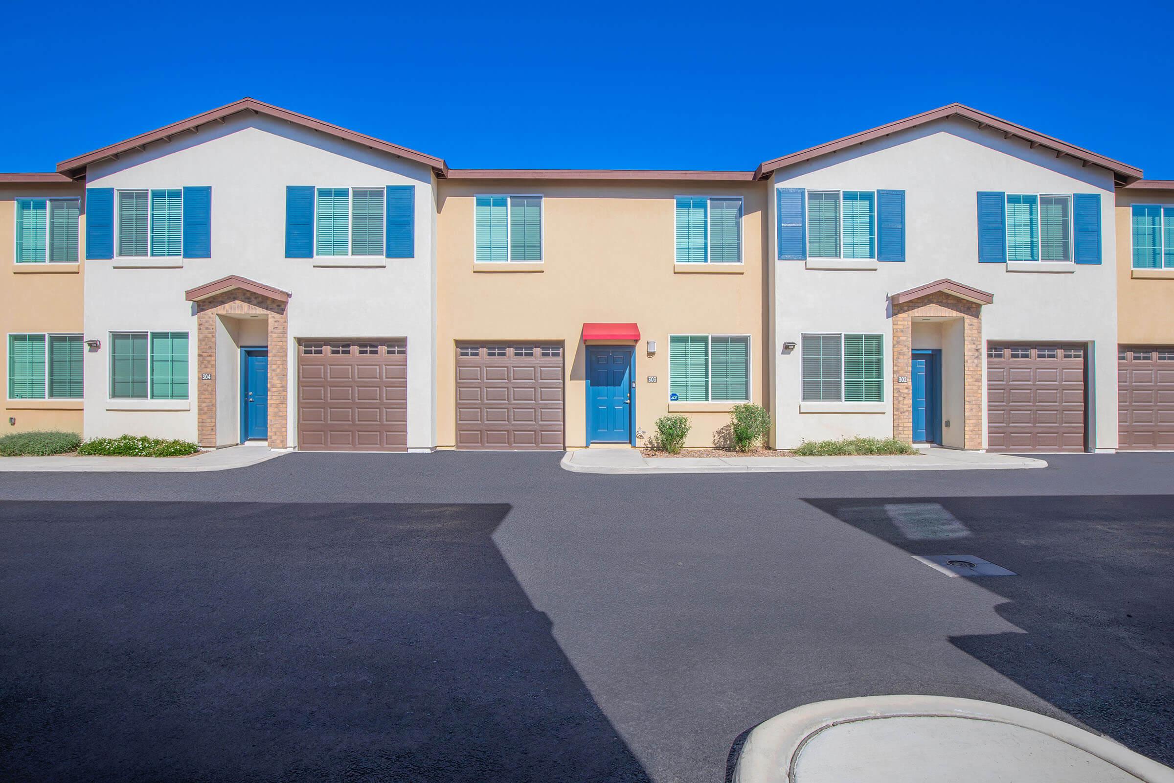 A row of modern townhouses with two stories, featuring blue shutters and doors. Each unit has a garage with brown doors. The sky is clear and blue, and the pathway is paved. The area is neatly landscaped with shrubs, creating a tidy residential environment.
