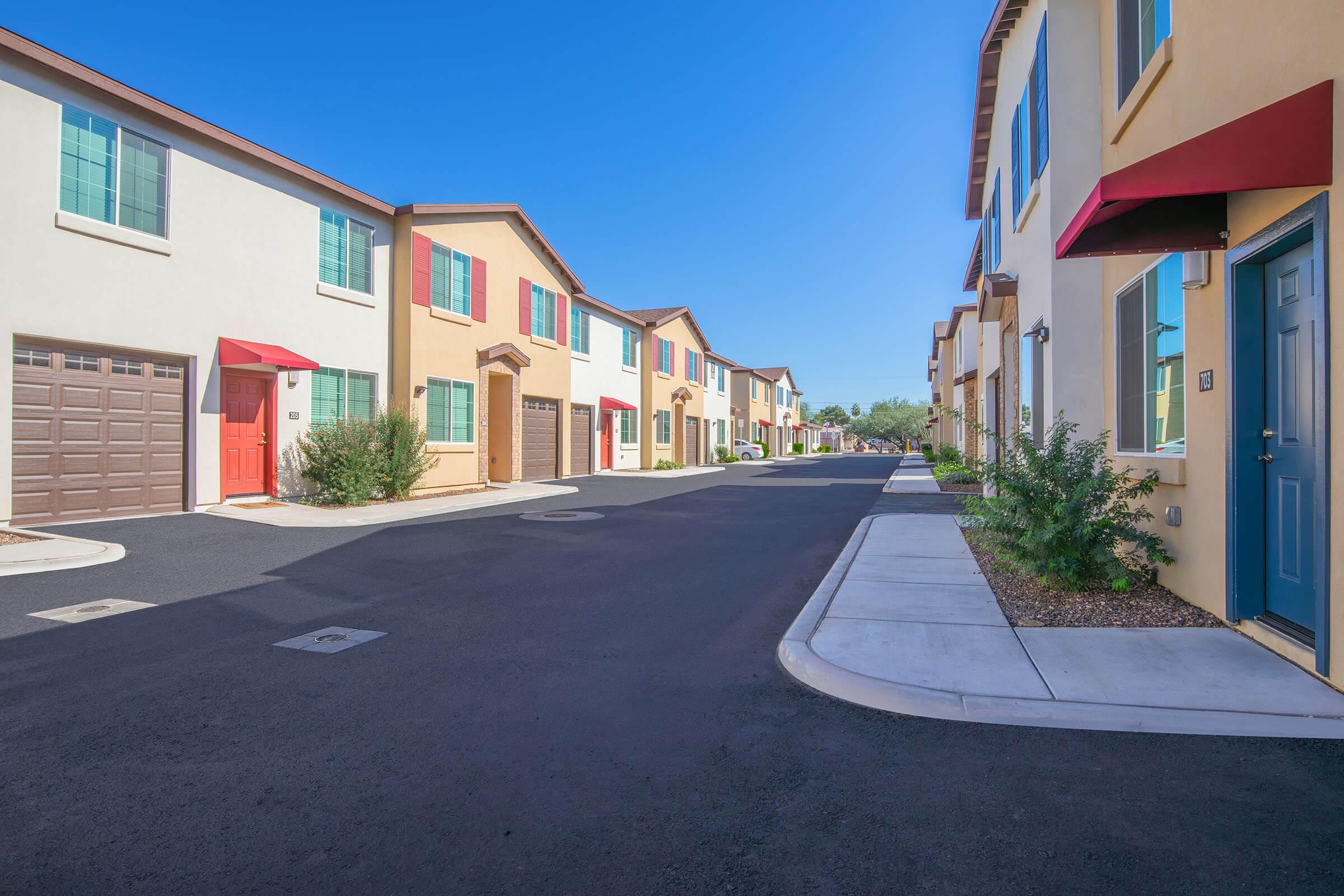 A residential street lined with colorful two-story townhouses. Each townhouse features a distinct entryway with windows, and some have awnings. The street is well-maintained, with a clear blue sky overhead and greenery along the sides, creating a vibrant and inviting atmosphere.