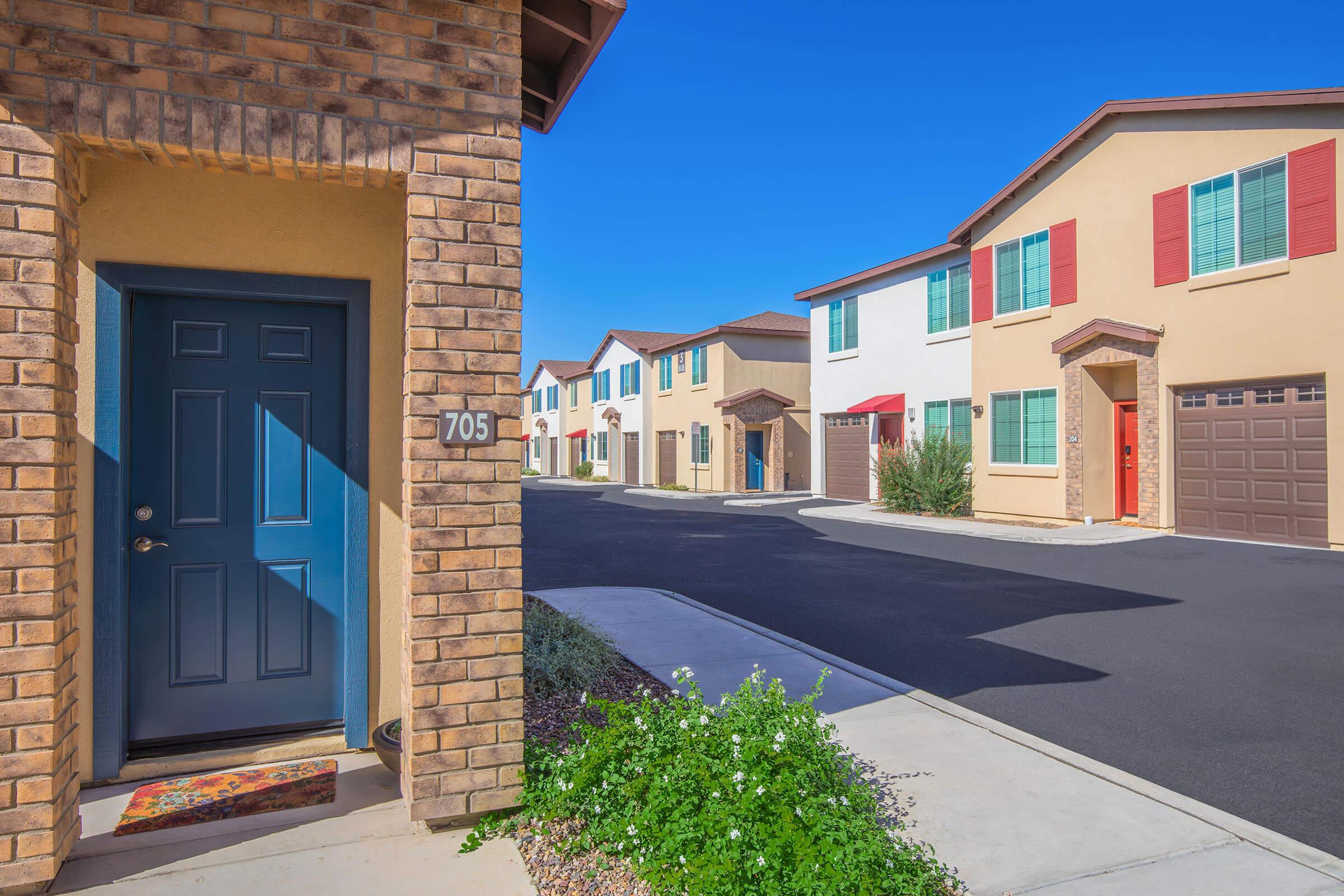A view of a residential neighborhood featuring a brick-built entrance with a blue door and the number 705 visible. The scene includes several colorful townhouses lining the street, with a clear blue sky above and a well-maintained sidewalk with greenery in front.