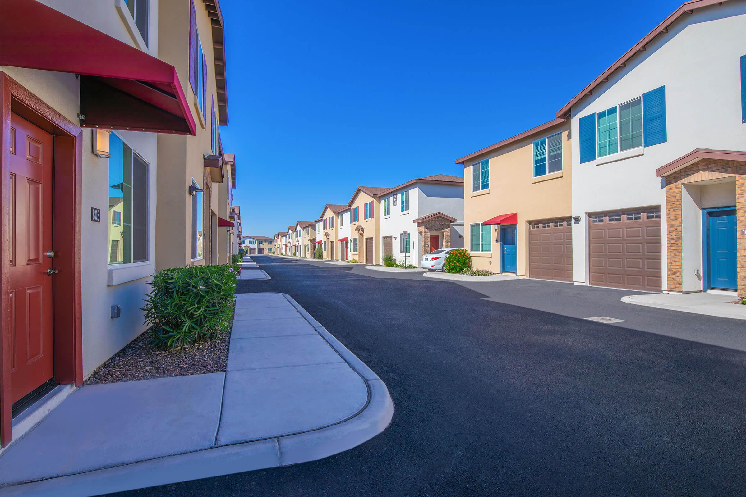 A picturesque residential street lined with modern townhouses featuring colorful facades. The view captures a clear blue sky, well-maintained sidewalks, and doorways with awnings. Some houses display garages, and neat landscaping adds to the inviting atmosphere of the community.