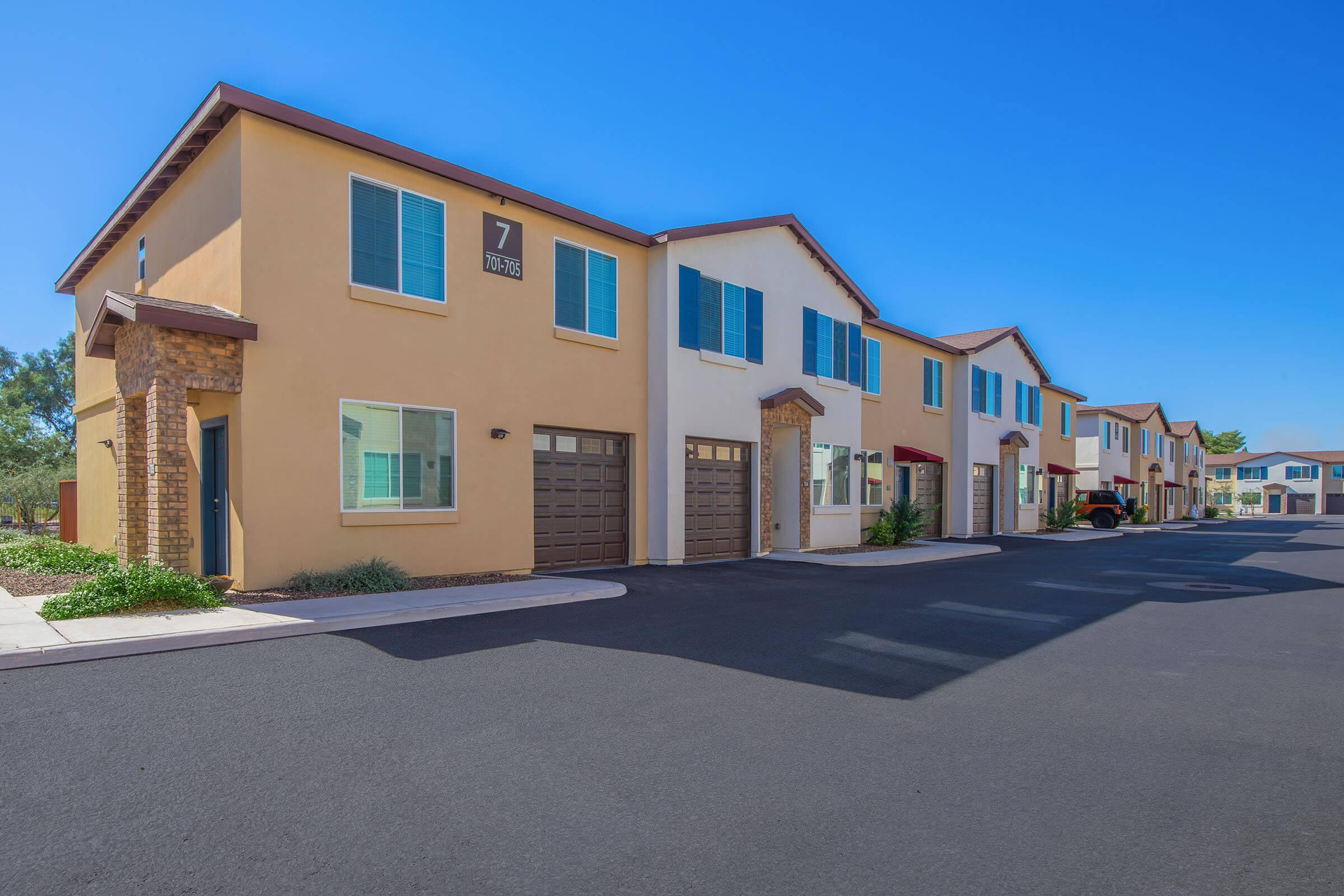 Row of modern, two-story townhouses with beige and brown exteriors, featuring garages and blue windows. The street in front is freshly paved and lined with small shrubs and plants. A clear blue sky provides a bright backdrop, creating a welcoming residential atmosphere.