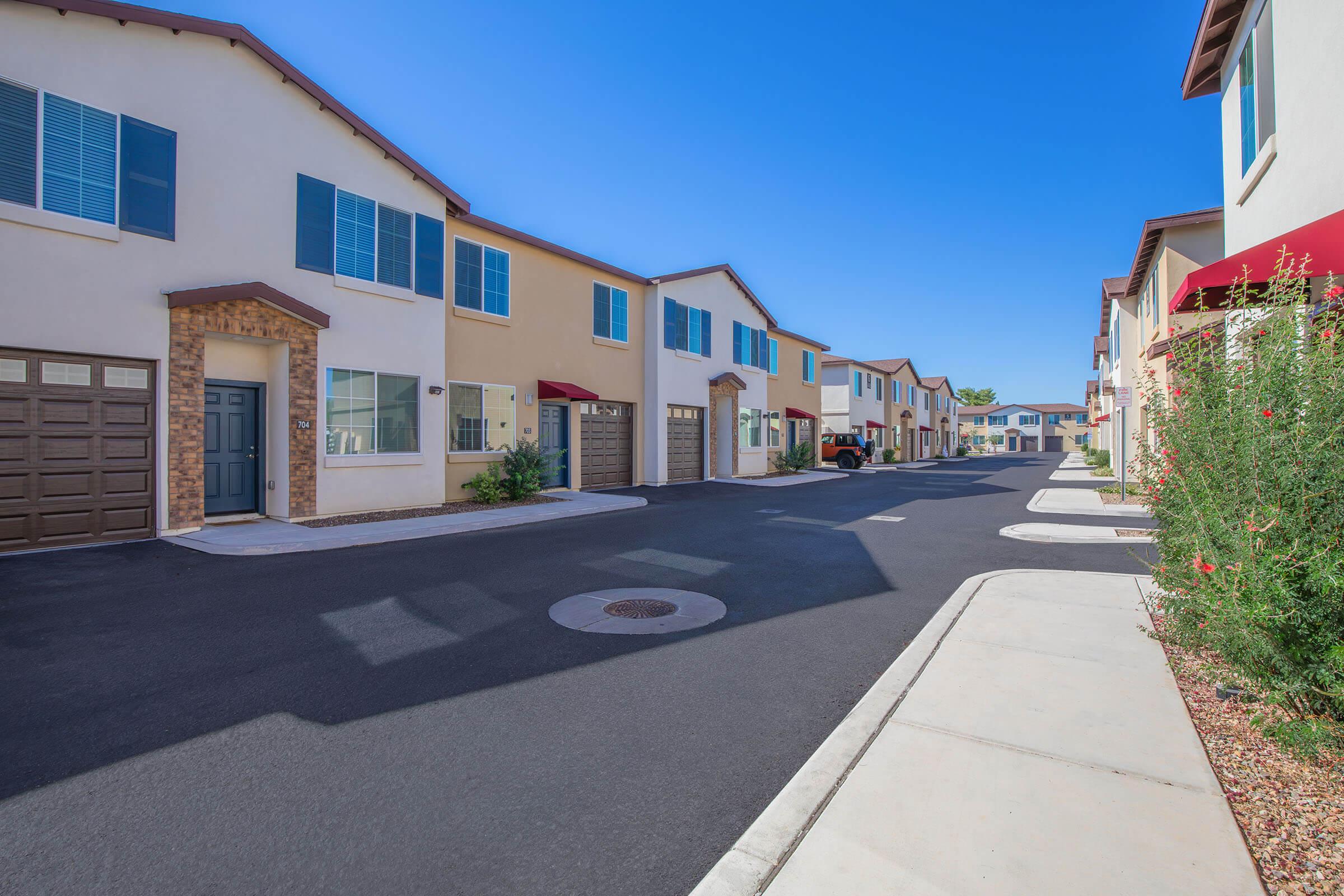 A peaceful residential street lined with modern two-story houses featuring beige and light blue exteriors. There are garages, manicured landscaping with colorful flowers, and a clear blue sky overhead. The street is clean, with well-maintained pavement and a manhole cover visible.
