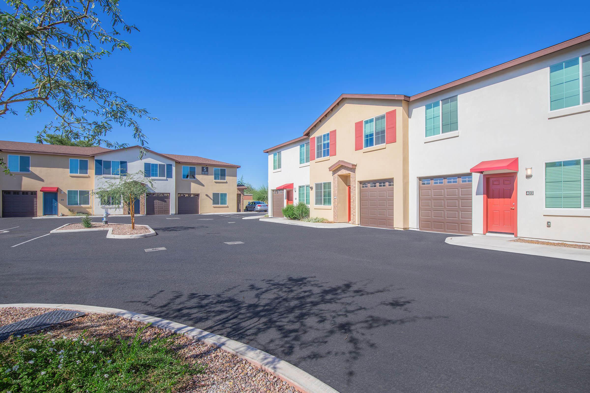 A modern residential complex featuring two-story houses with beige and red accents. The area includes paved parking spaces and landscaped greenery, set against a clear blue sky. There are multiple buildings arranged in a welcoming layout, creating a community atmosphere.