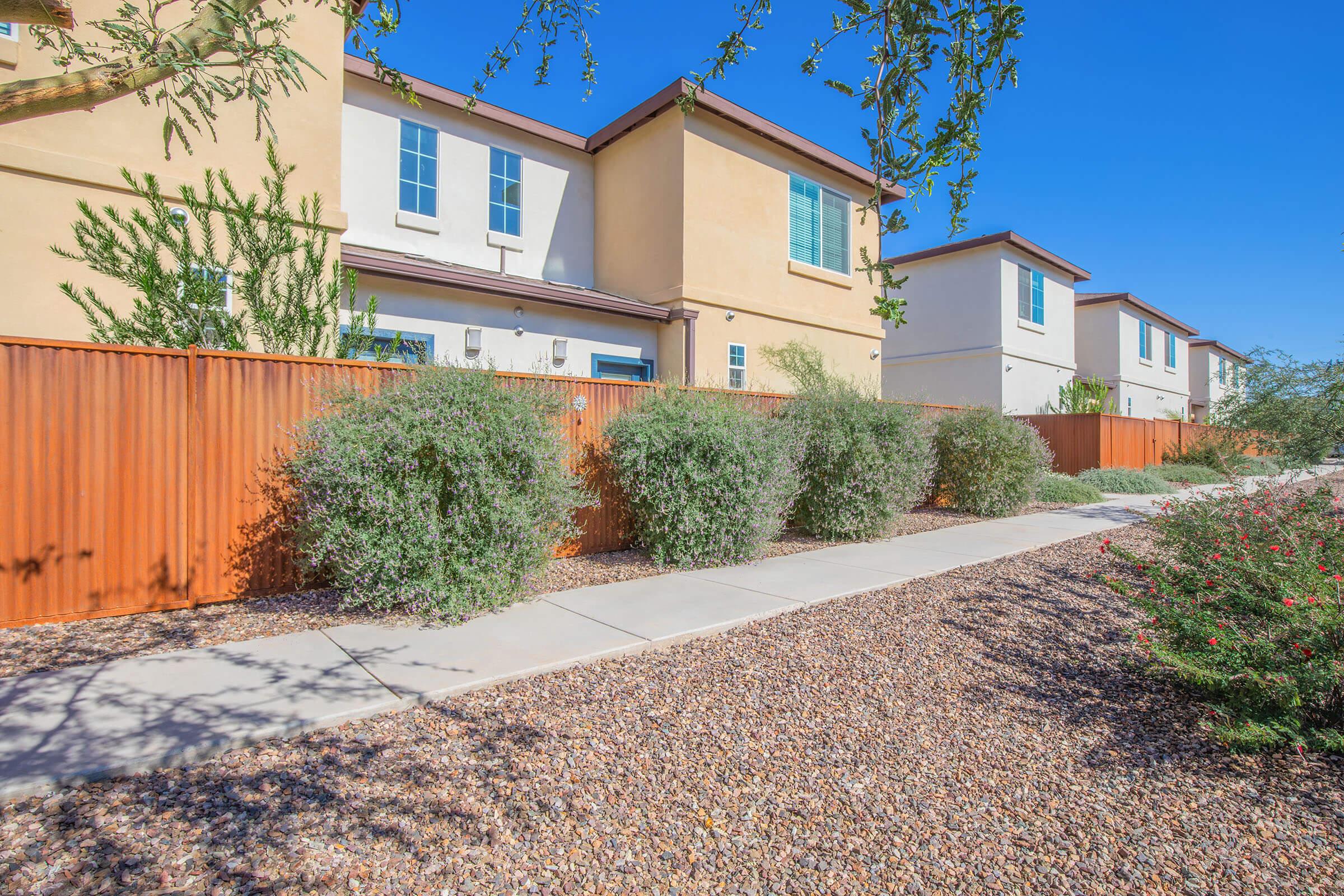 A row of residential buildings with beige exteriors and blue windows beside a landscaped pathway. Lush green shrubs and small trees line the path, while a rust-colored fence adds contrast. The sky is clear and blue, creating a bright and welcoming atmosphere.