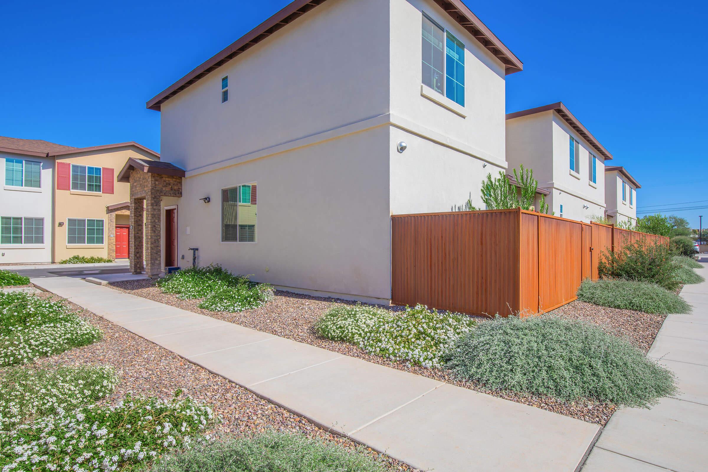 A modern residential building with a beige exterior and red door. The pathway is lined with small white flowers and green plants. A wooden fence is visible to the right, and blue skies enhance the bright, cheerful atmosphere of the neighborhood.