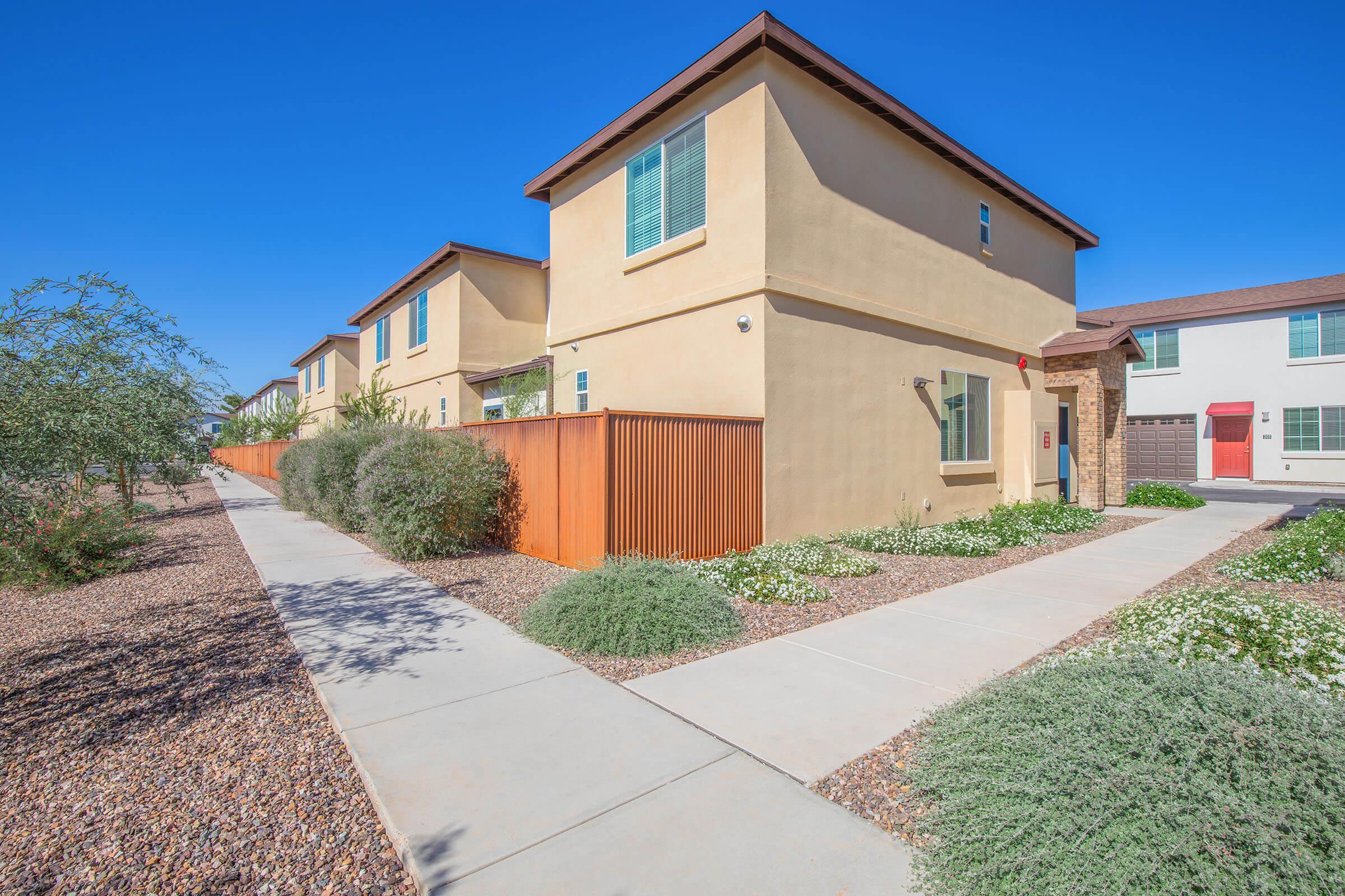 Two modern houses side by side on a sunny, clear day. Each house has a distinct facade, with one featuring light-colored stucco and large windows. The pathway is lined with small shrubs and flowers, and a wooden fence separates the front yards, creating a neat and inviting atmosphere.