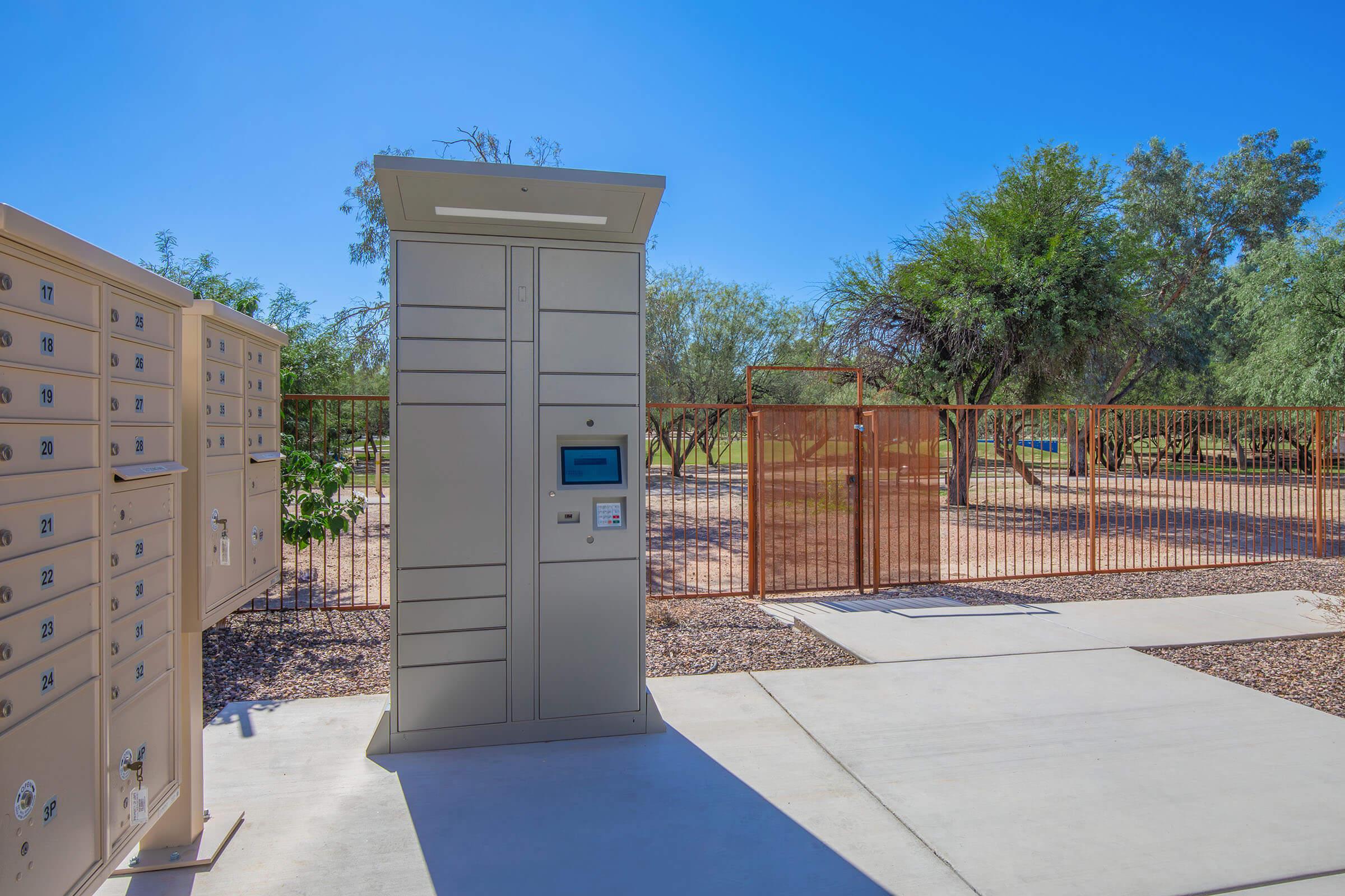 A modern package delivery locker system stands beside a row of mailboxes, set in a sunny outdoor area with sparse vegetation and a fenced backdrop. The scene features a clear blue sky and a concrete pathway leading to the lockers, providing convenient access for residents.