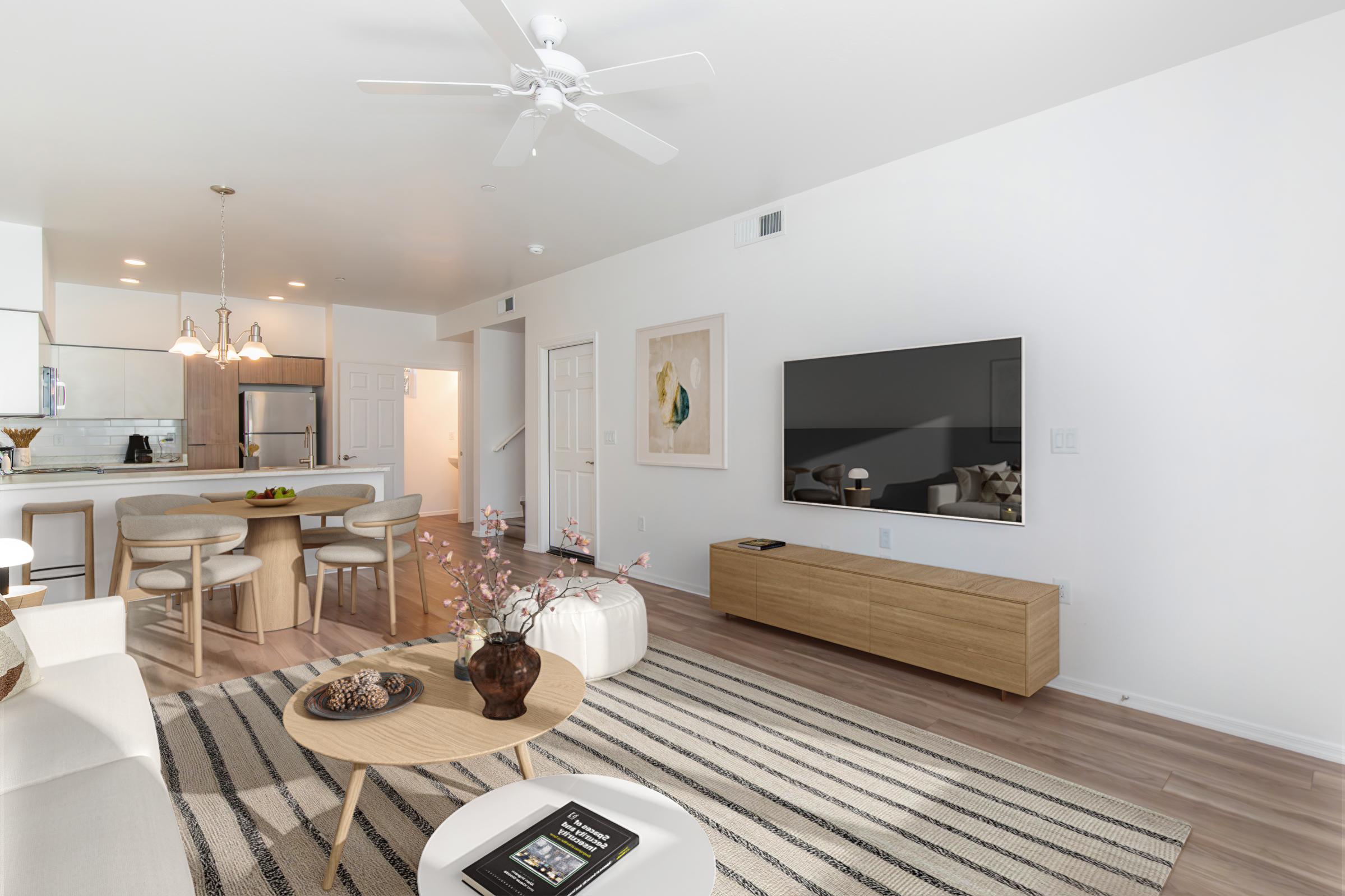 A modern living room featuring a light color palette with a large TV mounted on the wall and a wooden media console. A round coffee table sits in the center, adorned with decorative items. The space includes a cozy couch and an open kitchen area in the background, complemented by a dining table with chairs.