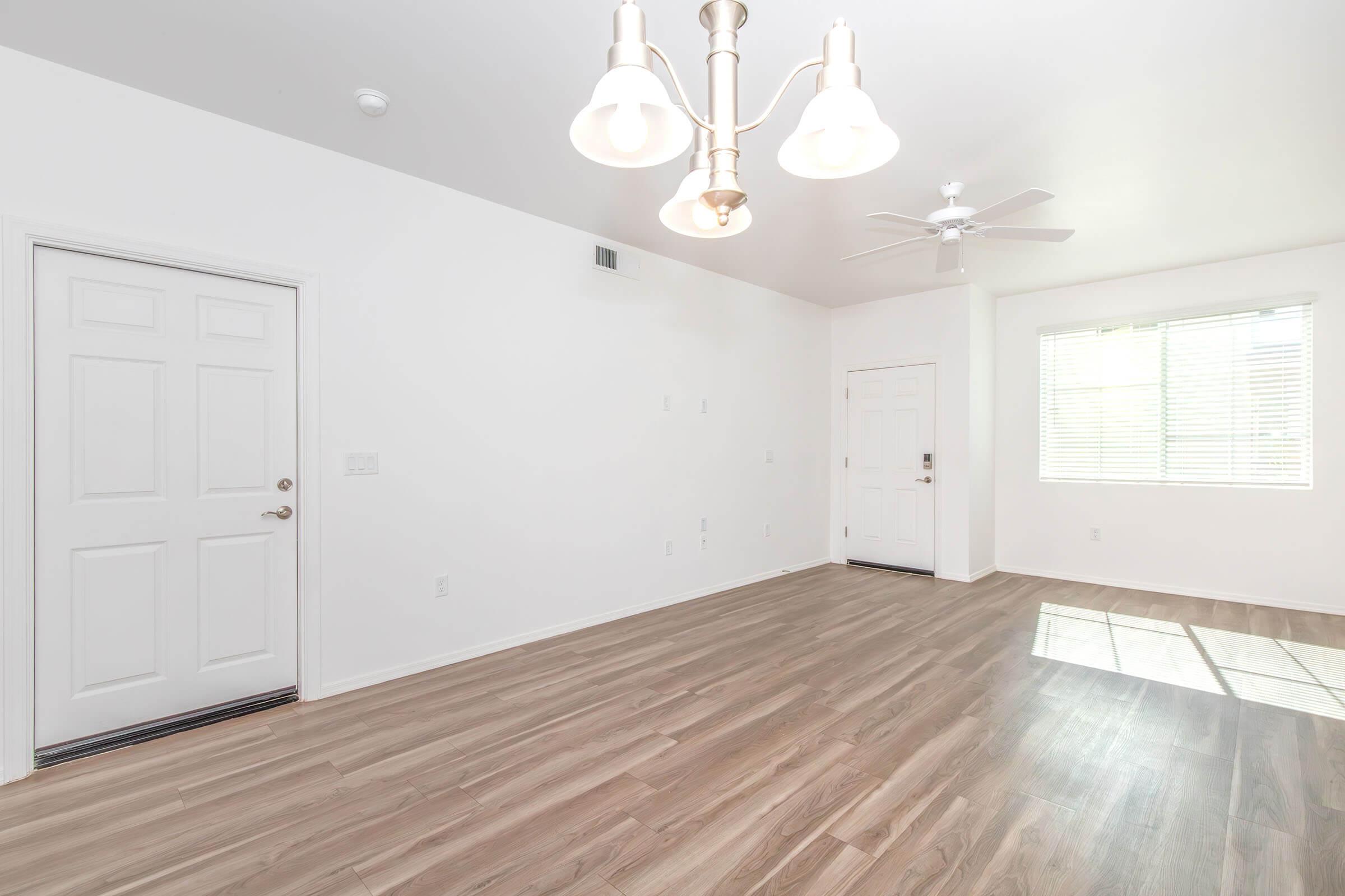 Empty room with light-colored walls and a wooden floor. There's a light fixture with three bulbs hanging from the ceiling, a ceiling fan, a window with blinds on the right side, and two doors: one leading outside and another to an adjoining room. Natural light is illuminating the space.