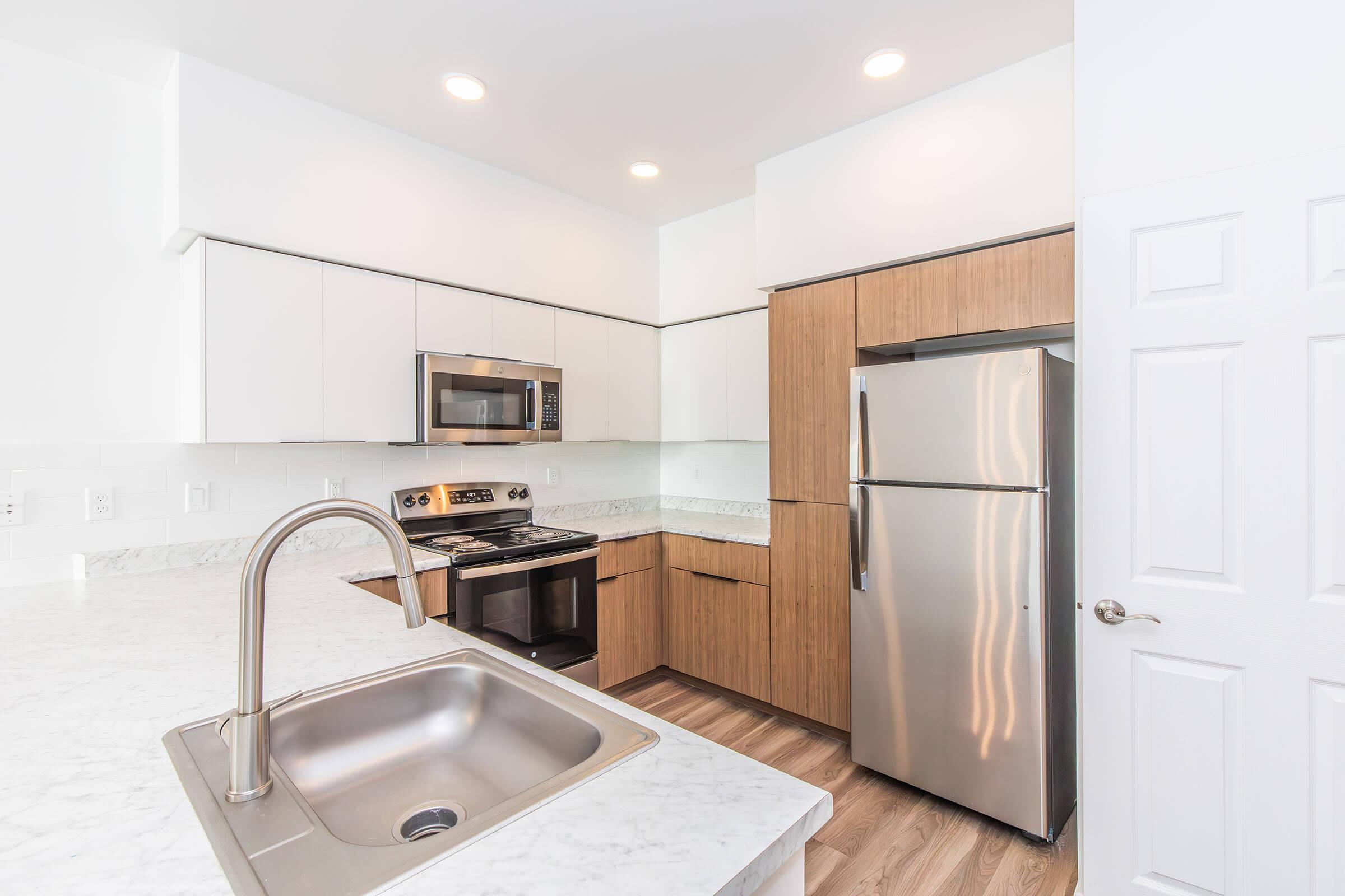 Modern kitchen featuring a marble countertop, stainless steel appliances including a microwave and oven, wooden cabinetry, and a sleek refrigerator. Bright, open space with natural light and a minimalist design. The sink is integrated into the countertop, enhancing the contemporary look.