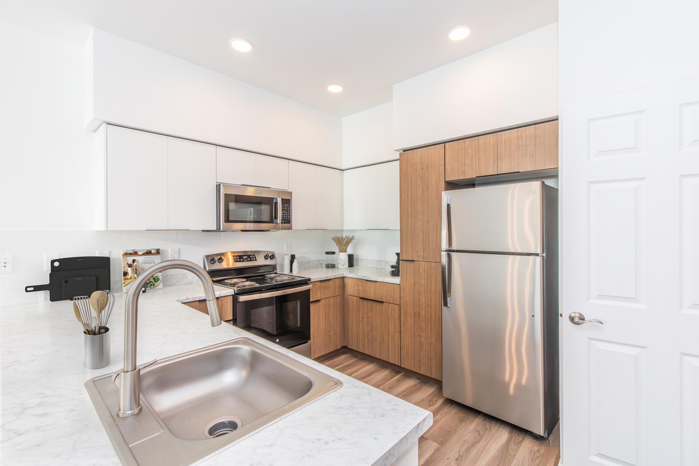 Modern kitchen featuring white cabinetry and wood accents. Includes stainless steel appliances such as a microwave and refrigerator, a marble countertop, and a sink. Bright lighting enhances the contemporary design, while a bowl of utensils adds a functional touch. Floor is made of light wood.