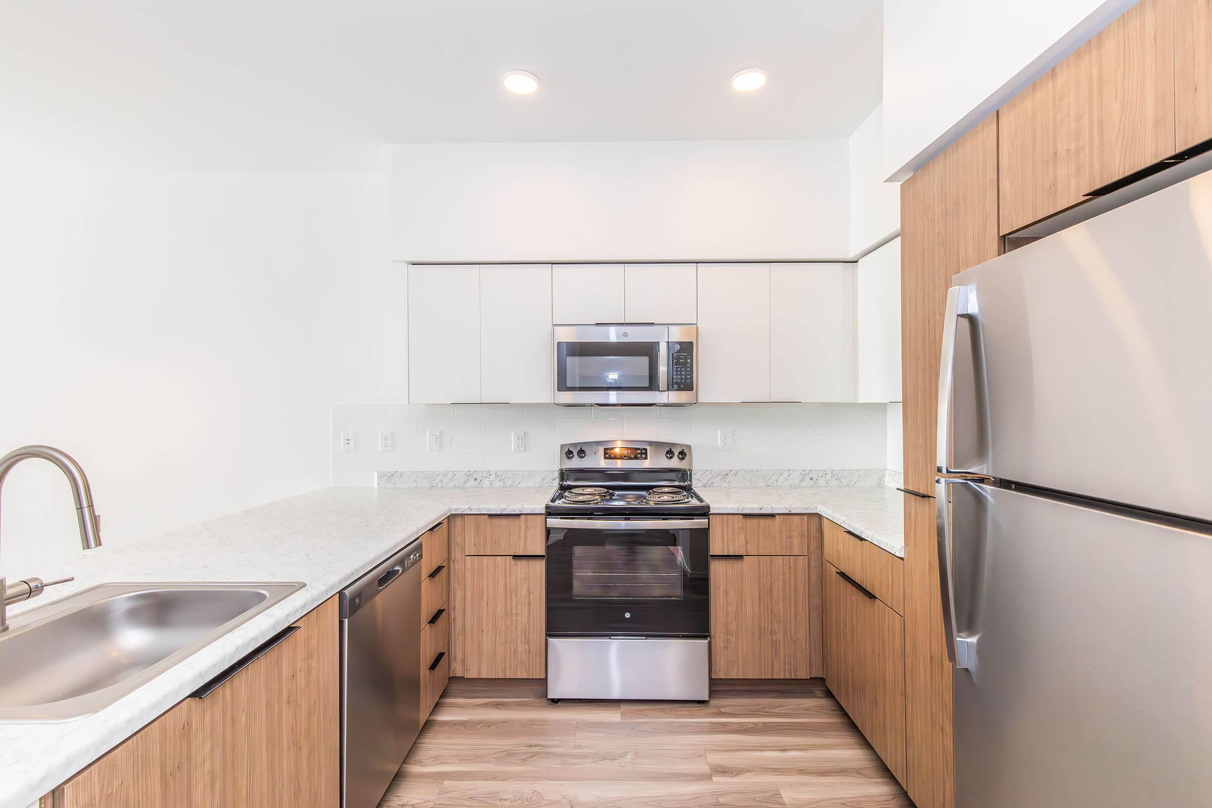Modern kitchen featuring light wood cabinetry, stainless steel appliances including a microwave and oven, a double basin sink, and a white countertop. The space is bright with recessed ceiling lighting and a clean, minimalist design, showcasing an open layout.