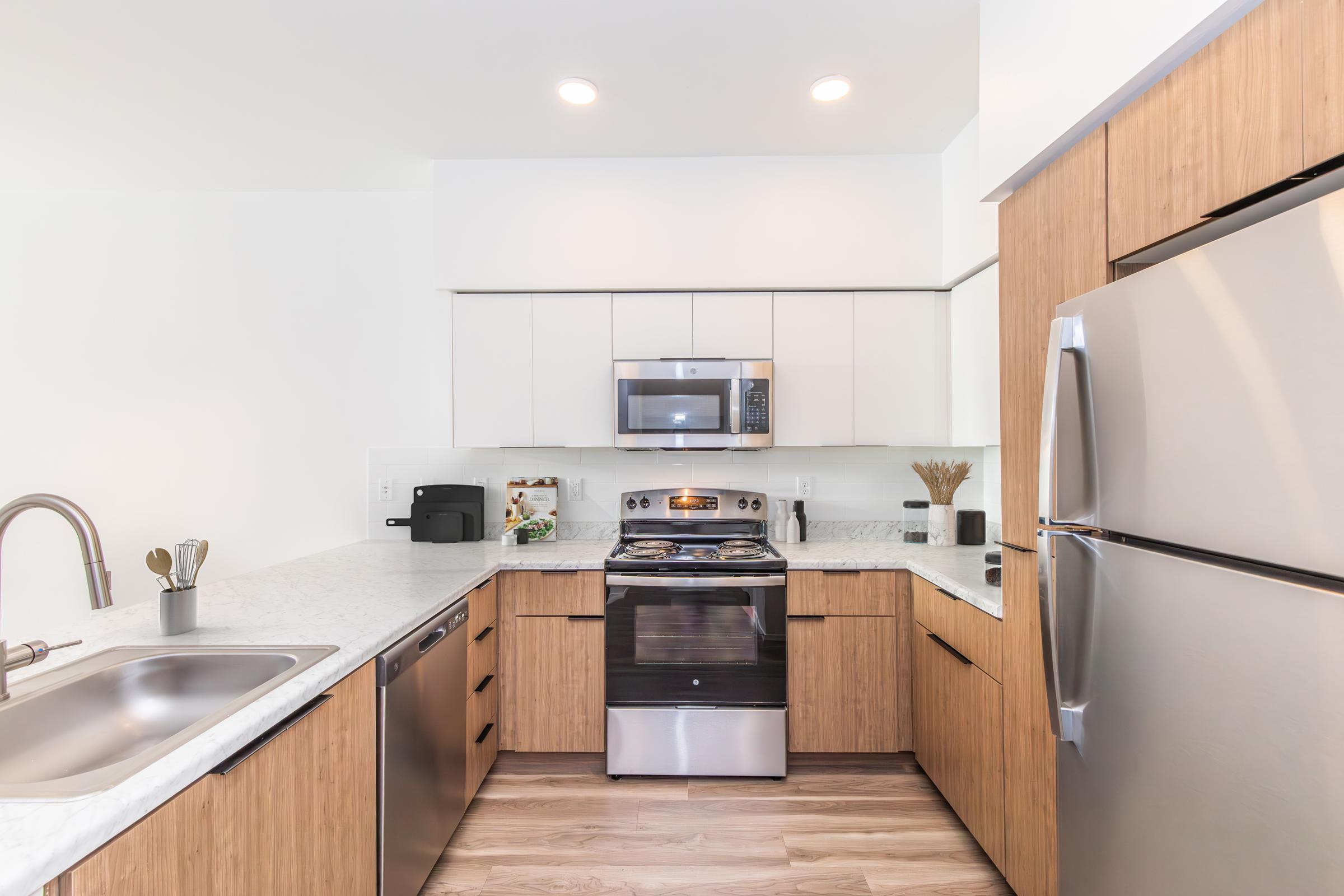 Modern kitchen featuring wooden cabinetry, marble countertops, a stainless steel refrigerator, and built-in microwave above a gas stove. The sink is positioned on the left, with kitchen utensils visible. Soft lighting illuminates the space, creating a bright and inviting atmosphere.