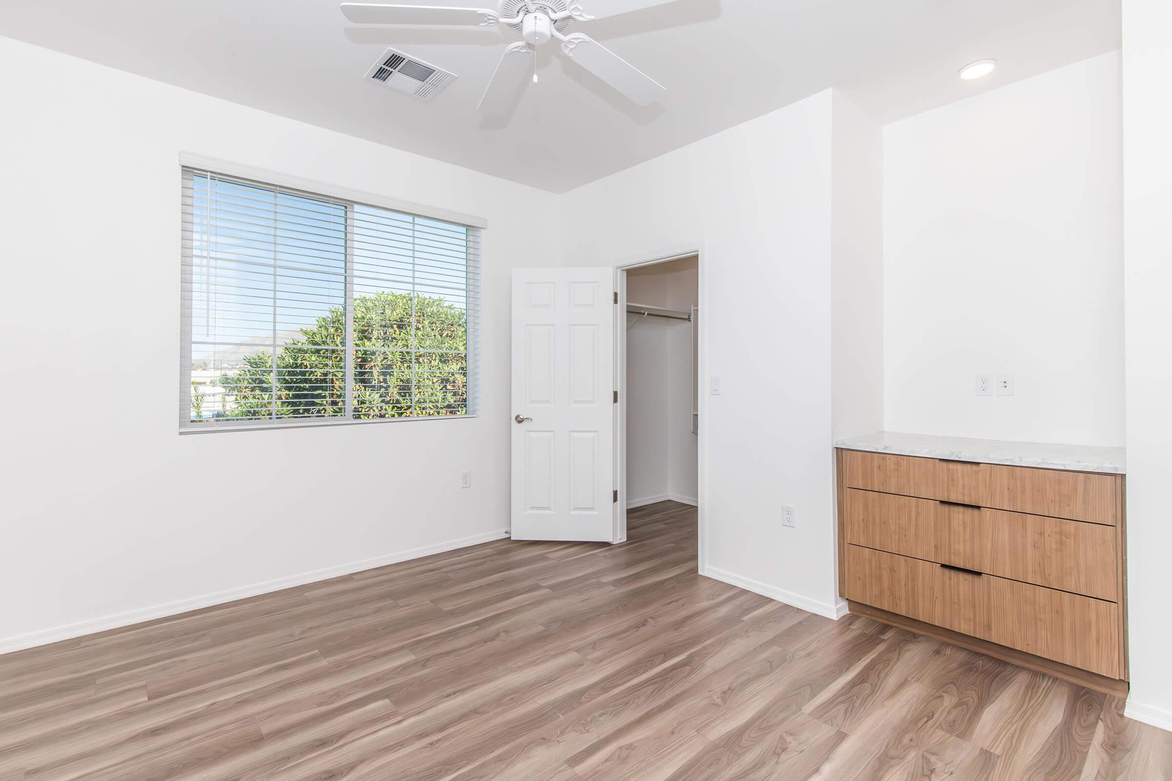 A bright, unfurnished room featuring light hardwood flooring, a ceiling fan, and a window with blinds. A closet is visible through an open door, and a cabinet with wooden drawers is located near the wall. The walls are painted white, creating an airy and spacious atmosphere.