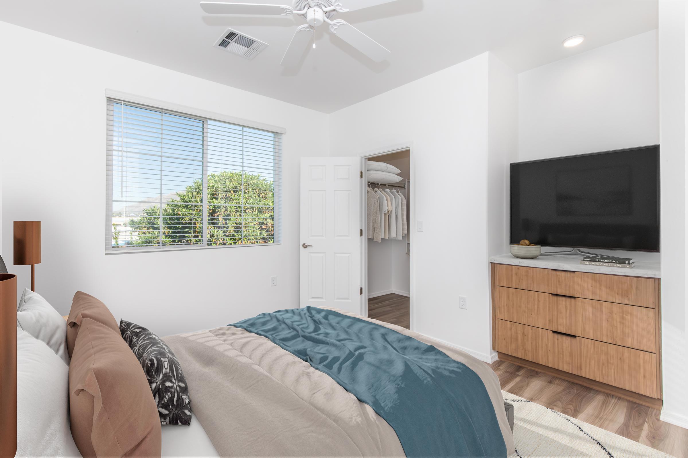 A modern bedroom featuring a neatly made bed with decorative pillows, a dresser, and a wall-mounted television. A window with blinds offers natural light and a view of greenery outside. A door leads to a closet, complementing the clean, minimalist design.