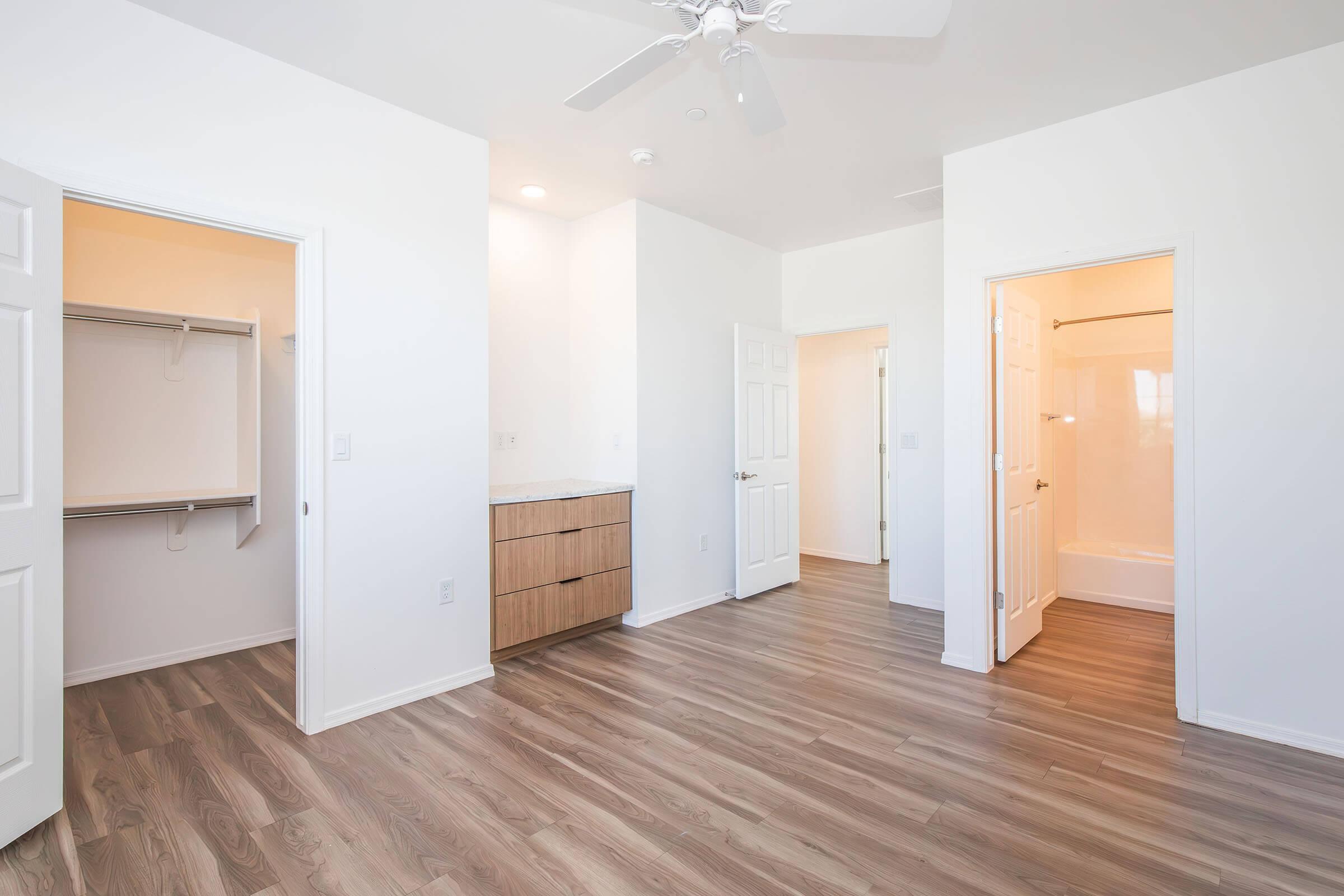 A spacious, unfurnished room featuring light wood flooring, white walls, and a ceiling fan. There are two open doors leading to a closet and a bathroom area. Natural light brightens the space, creating a clean and modern atmosphere suitable for various uses.