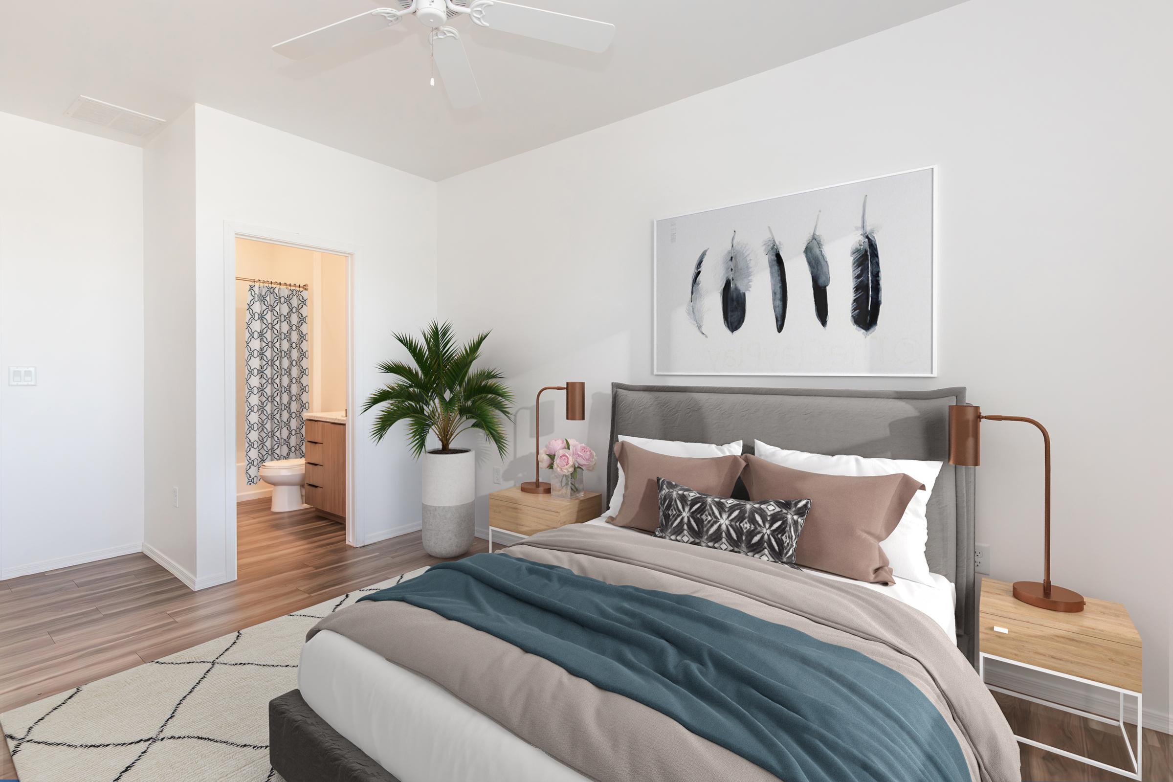 A modern, bright bedroom featuring a gray upholstered bed with plush pillows, a cozy blanket, and wooden bedside tables with lamps. A decorative feather artwork hangs above the bed. In the background, there is a doorway leading to a bathroom, and a potted plant adds a touch of greenery to the room.