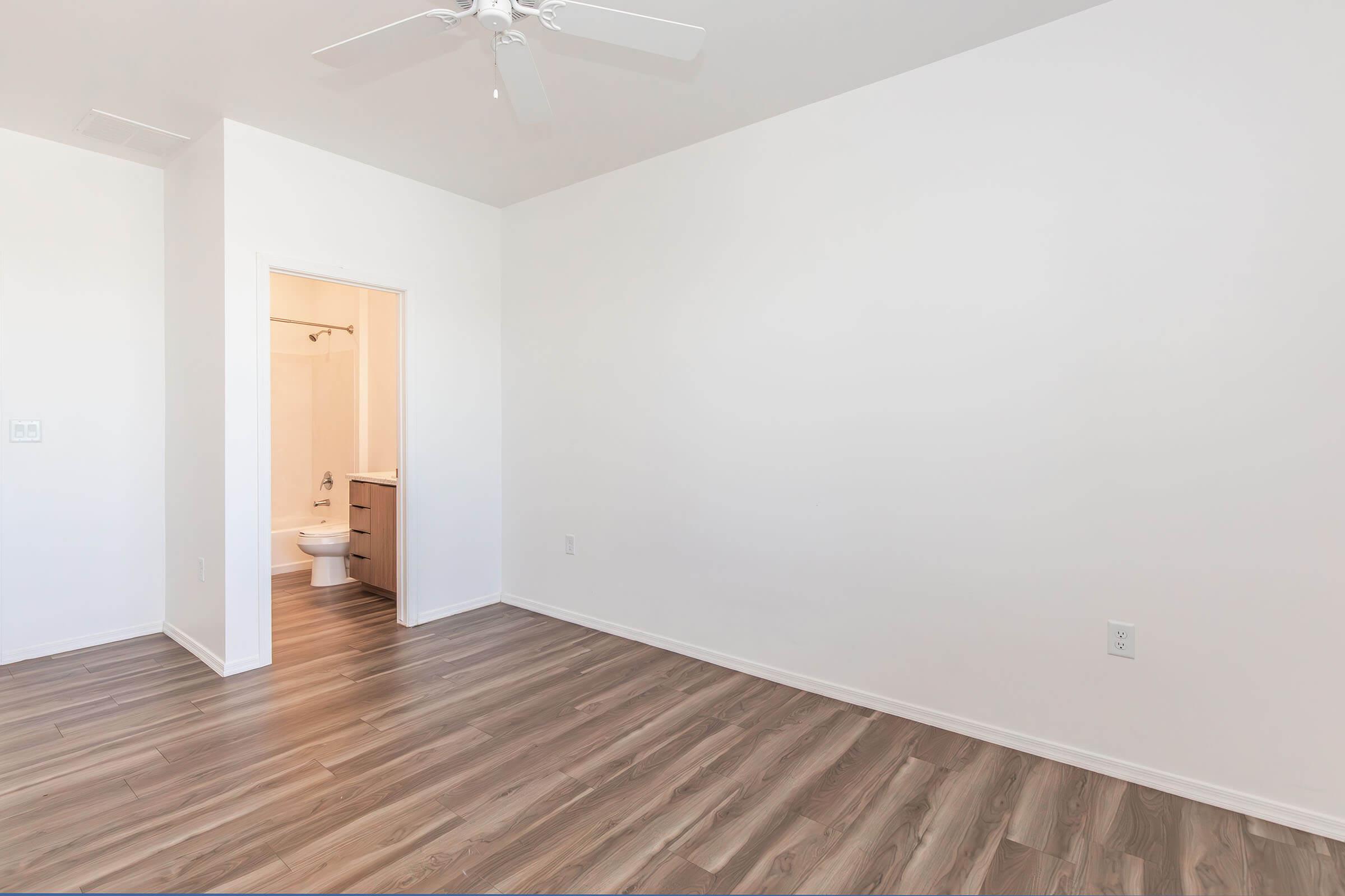 A spacious, empty room with light-colored walls and wood-like flooring. A door on the left leads to a bathroom, visible through the open doorframe. The ceiling features a ceiling fan, and the overall lighting is bright and clean, giving a fresh and modern appearance to the space.