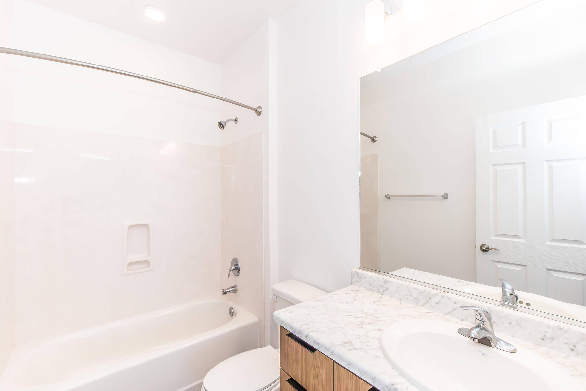 A clean and modern bathroom featuring a bathtub with a shower curtain, a marble countertop with a sink, a mirror above the sink, and white walls. A towel rack is mounted beside the mirror, and a closed bathroom door is visible in the background. The overall design is bright and minimalist.
