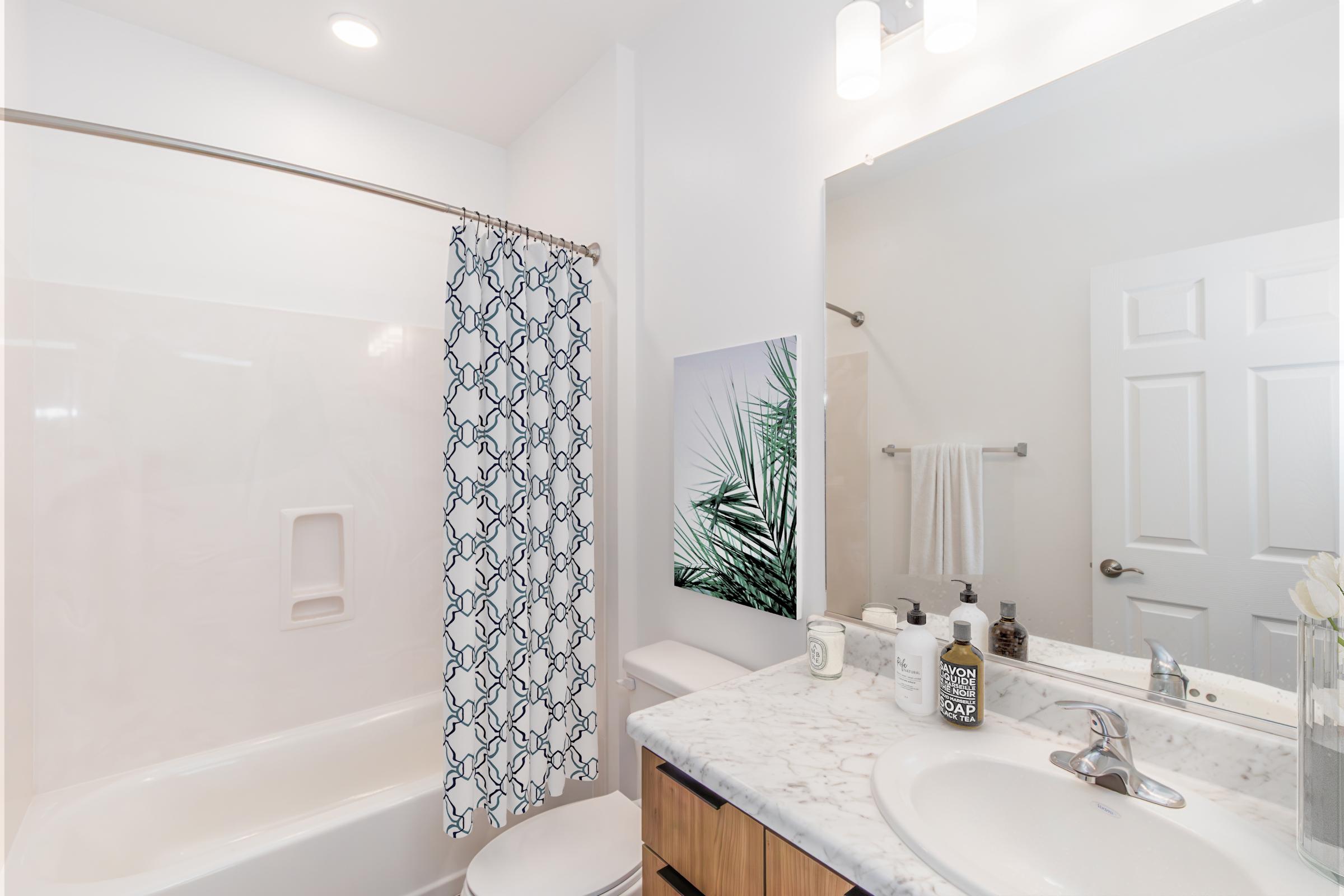A modern bathroom featuring a white bathtub with a shower, a blue and white patterned shower curtain, a marble countertop with various toiletries, a large mirror, and a plant-themed wall art. The space is well-lit with overhead lighting.