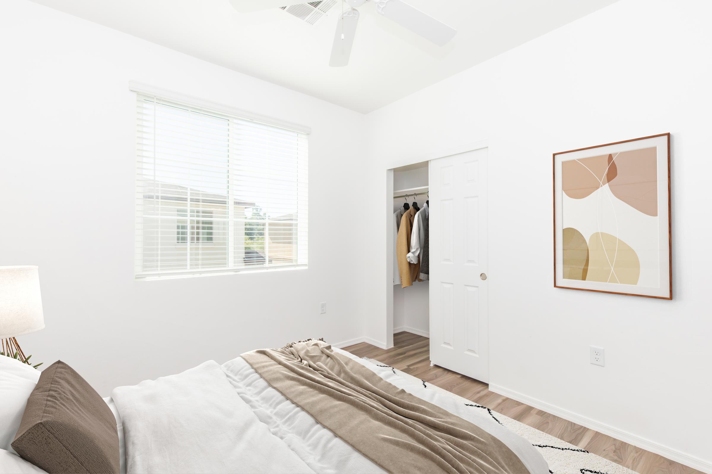 Bright and airy bedroom with a large window featuring white blinds, showcasing a neatly made bed with beige and white bedding. A cozy lamp on a side table, an artwork with abstract shapes on the wall, and a closet with clothes visible create a welcoming atmosphere. The room has light-colored walls and a simple rug.