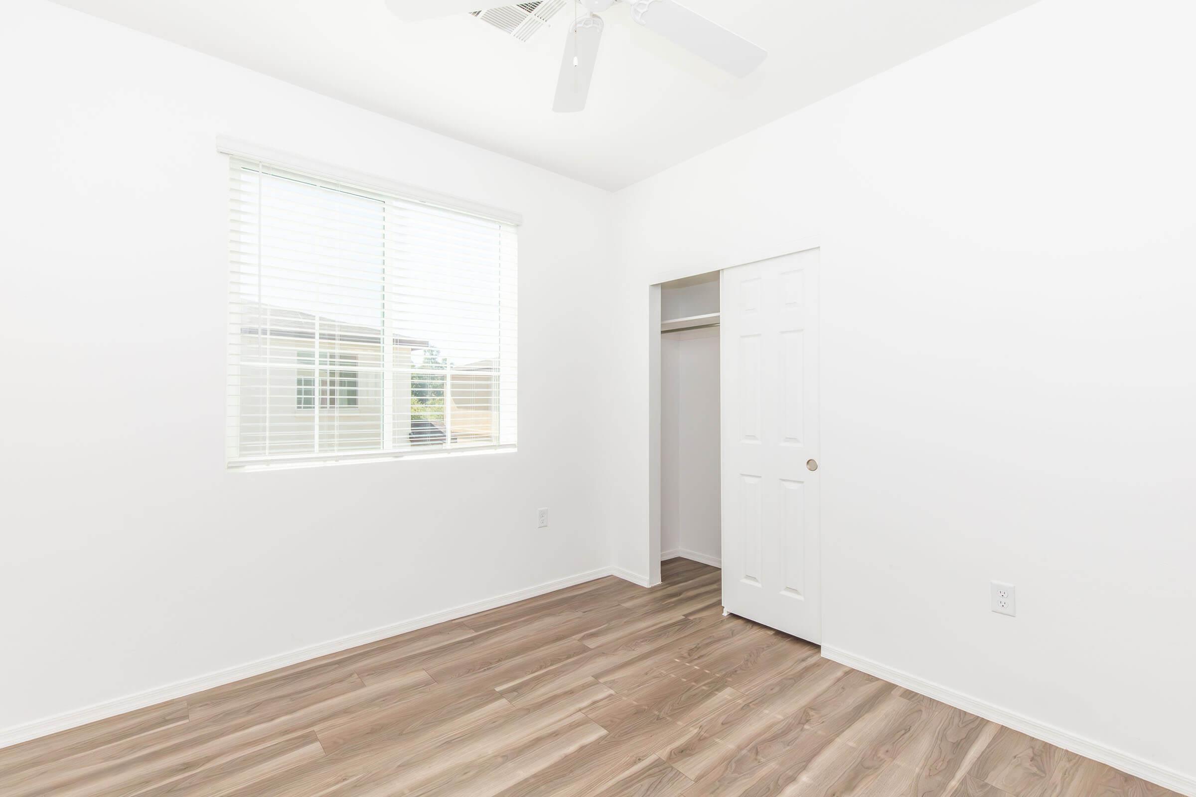 A bright, empty room with white walls and a window featuring blinds. There is a closet with a white door on the left, and the floor has light wood-like flooring. The ceiling has a ceiling fan, creating a clean and open atmosphere.