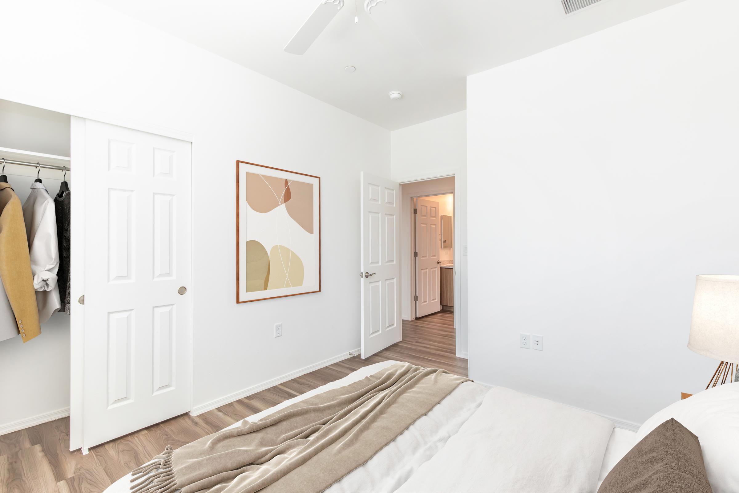 A modern bedroom featuring a neatly made bed with a beige and white striped throw blanket. A framed abstract art piece hangs on the wall. There are two white closet doors and an open doorway leading to a bathroom. The flooring is light wood, and the walls are painted white, creating a bright and airy atmosphere.