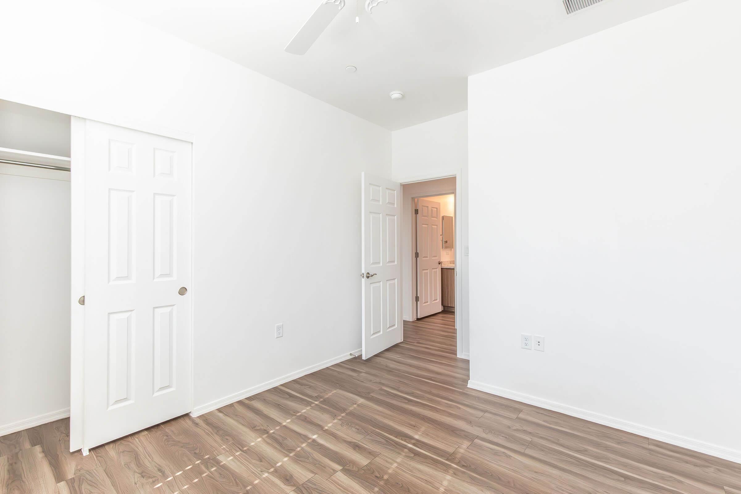 A spacious, empty room with white walls and a wooden floor. Two doors are visible: one leading to a closet and the other to a bathroom. Natural light streams in, creating a bright and open atmosphere. The room is designed for potential personalization and furnishing.