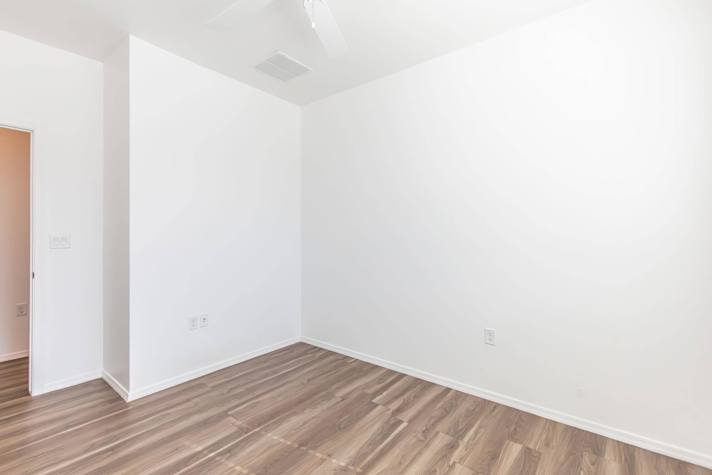 A bright, empty room with white walls and a wooden floor. There is a ceiling fan visible, and one wall features a doorway leading to another space. The room is well-lit and appears clean and spacious, suitable for various uses such as an office or bedroom.
