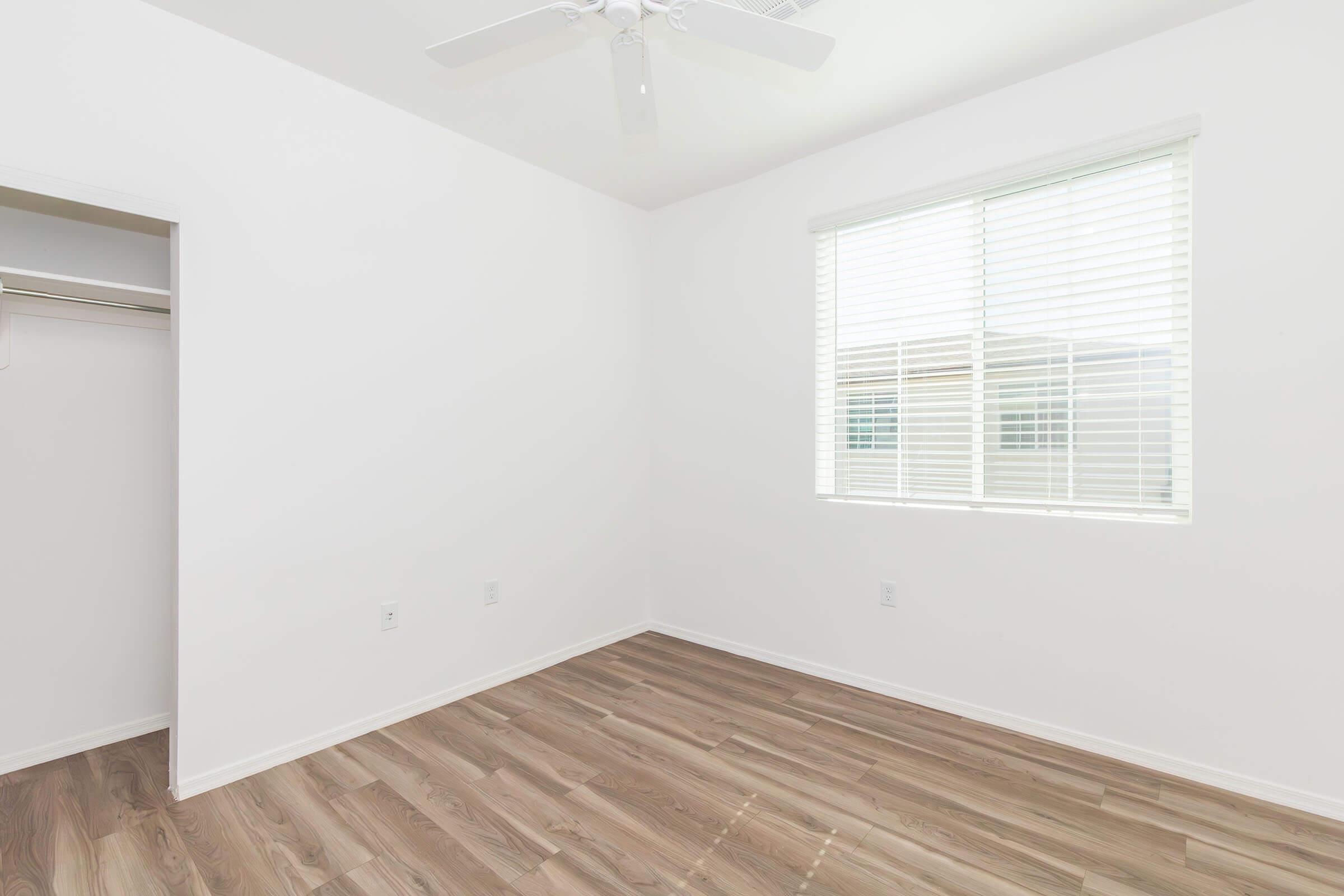 A bright and empty room with white walls and a light wooden floor. There is a window with blinds allowing natural light to enter. A ceiling fan is visible, and there is a small closet on one side. The overall atmosphere is clean and minimalist.