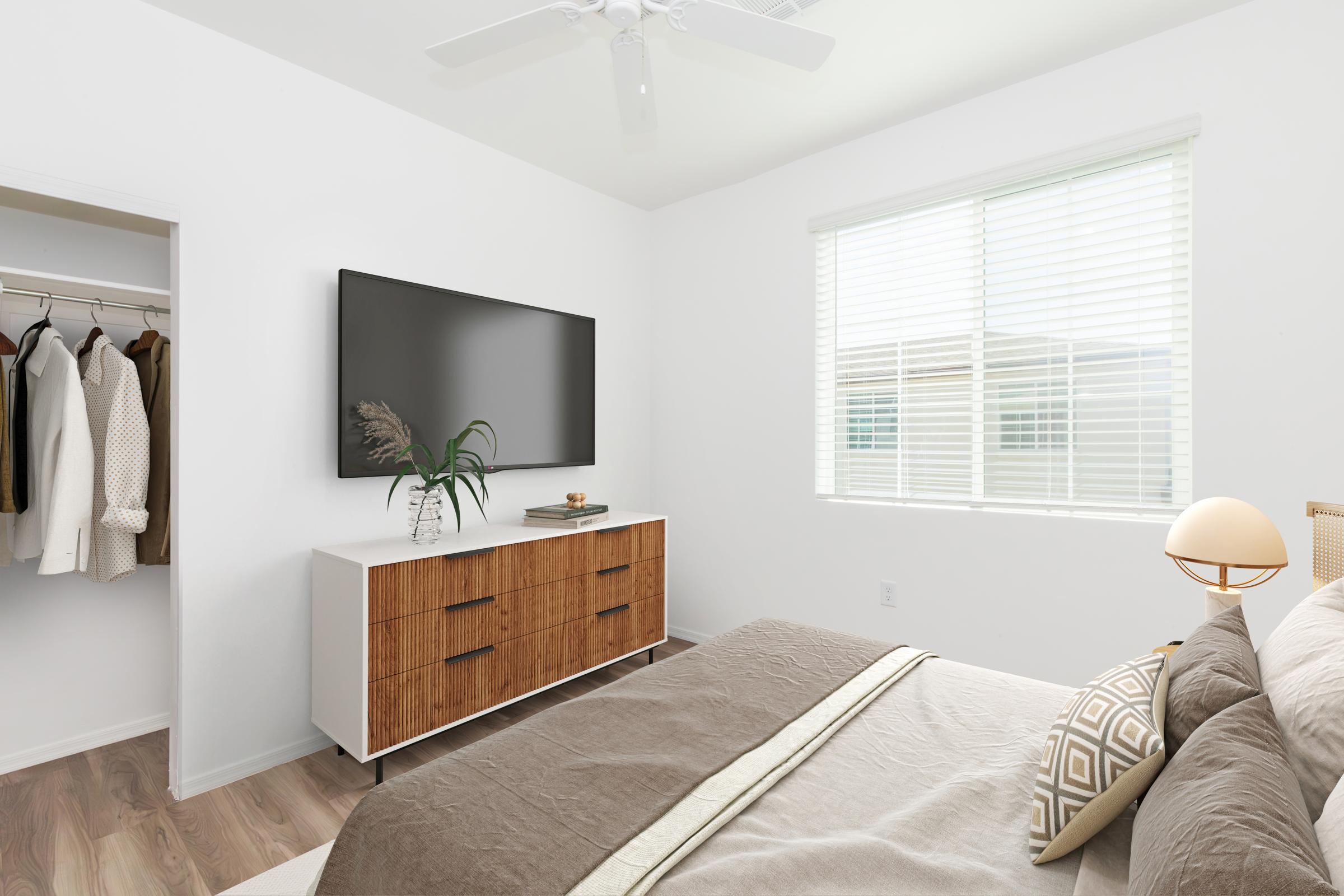 A modern bedroom featuring a neatly made bed with a neutral-colored blanket and decorative pillows. A stylish dresser with a plant and decorative items stands opposite a wall-mounted TV. A closet is visible on the left, and natural light streams in through a window with white blinds. The walls are painted white, creating a fresh look.