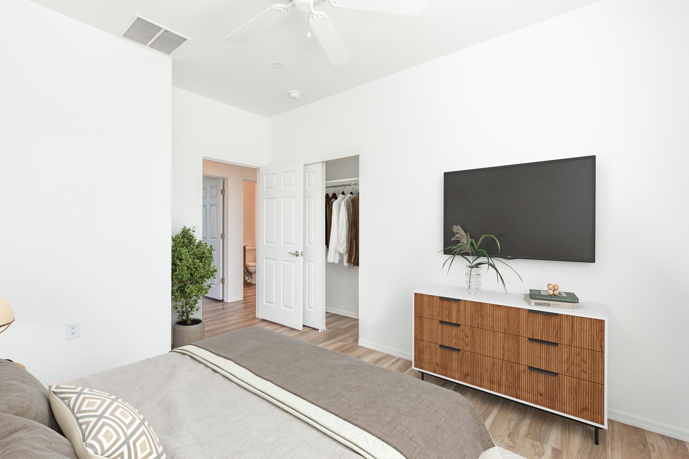 Modern bedroom featuring a cozy bed with a decorative throw, a wooden dresser with plants on top, and a wall-mounted TV. A closet with clothes is visible through an open door, and there's a small indoor plant adding a touch of greenery to the space. The walls are painted white, enhancing the bright atmosphere.