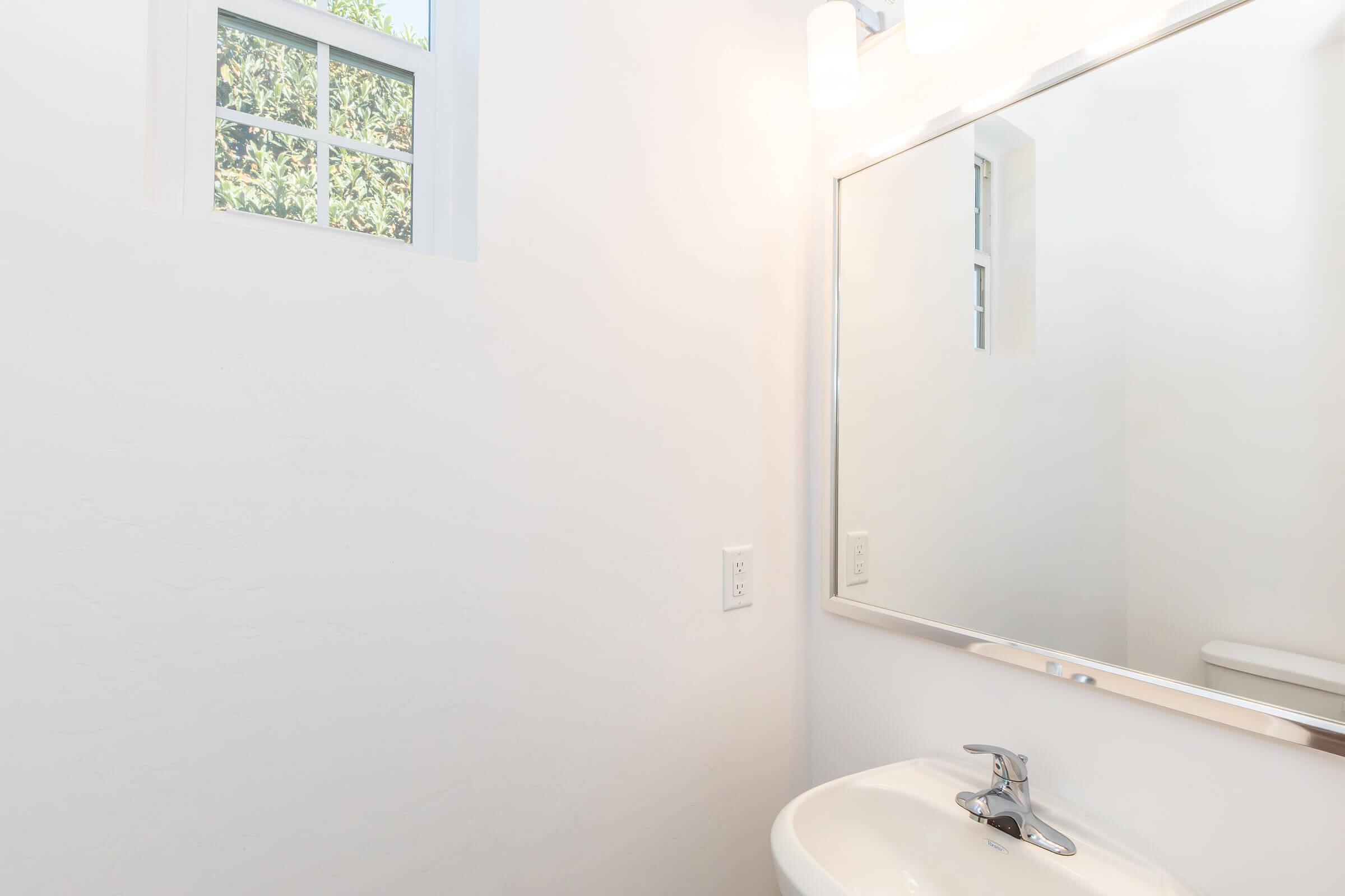 A bright, modern bathroom featuring a white wall and sink. A rectangular mirror is mounted above the sink, reflecting natural light from a nearby window. The overall design is minimalist and clean, creating an open atmosphere.