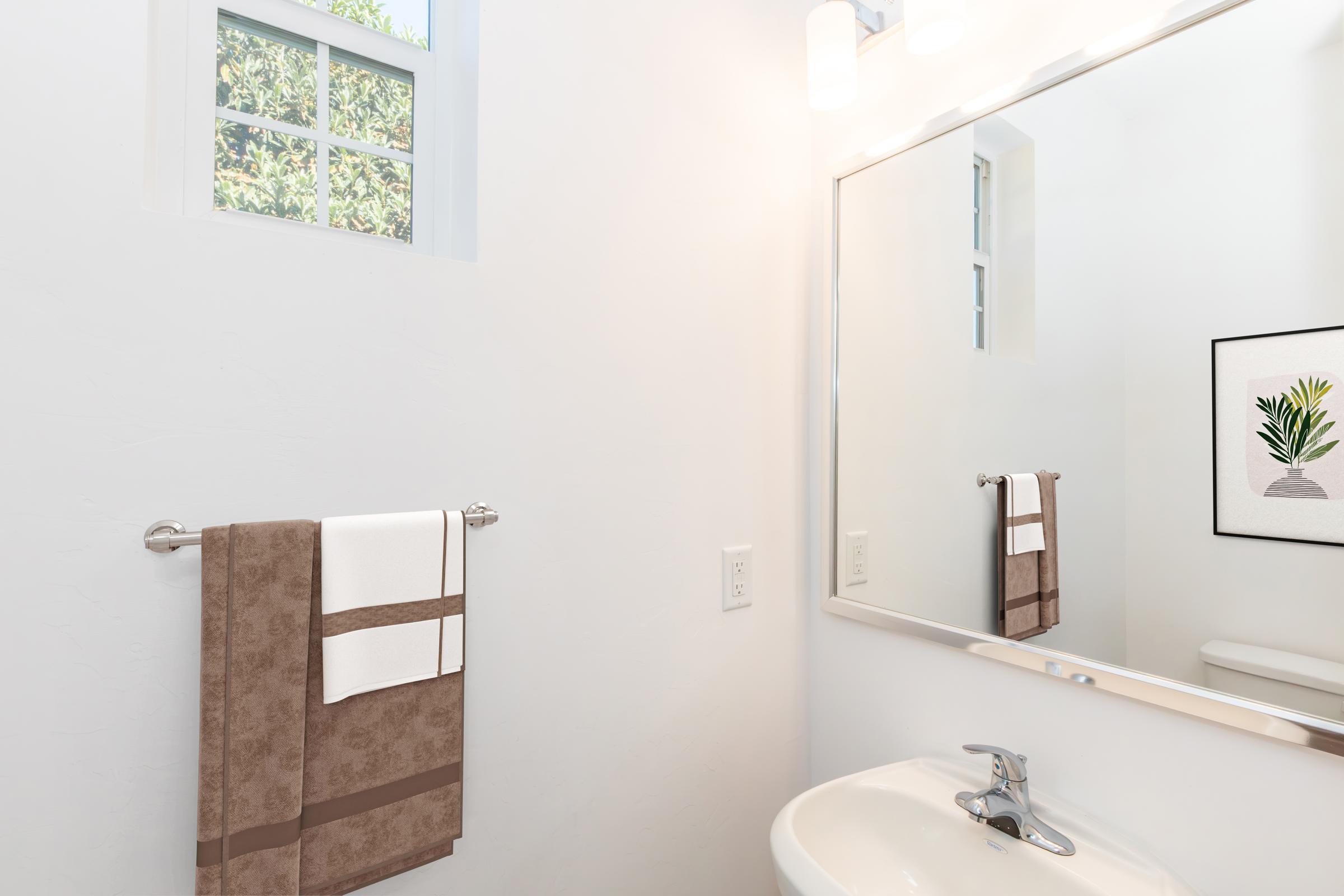 A bright and modern bathroom featuring a white wall, a large mirror, and a sink. There are two neatly arranged hand towels on a towel rack, and a small decorative plant print is framed on the wall in the background. A window lets in natural light, enhancing the airy feel of the space.