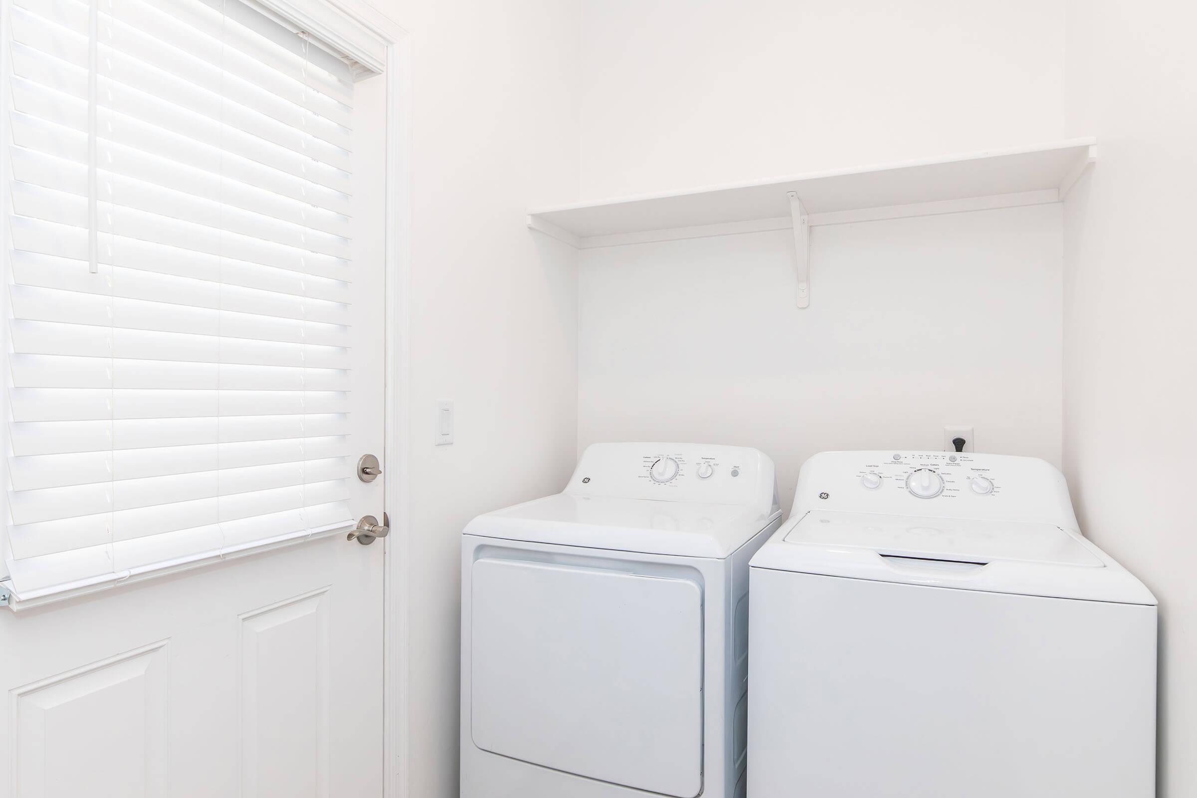 A bright laundry room featuring two white washing machines side by side against a white wall. There is a shelf above the machines and a door with a silver handle leading outside, complemented by a window with white blinds allowing natural light to enter the space.