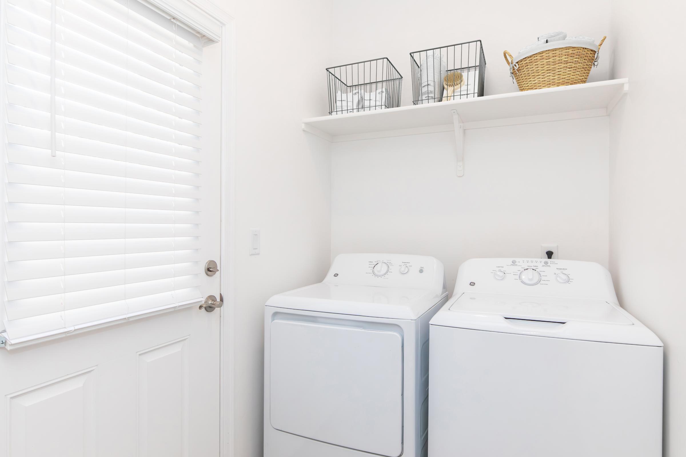 A clean and bright laundry room featuring a white washing machine and dryer side by side, with a shelf above holding black wire baskets and a wicker basket. There is a door with vertical blinds beside the appliances, enhancing the minimalistic and organized look of the space.