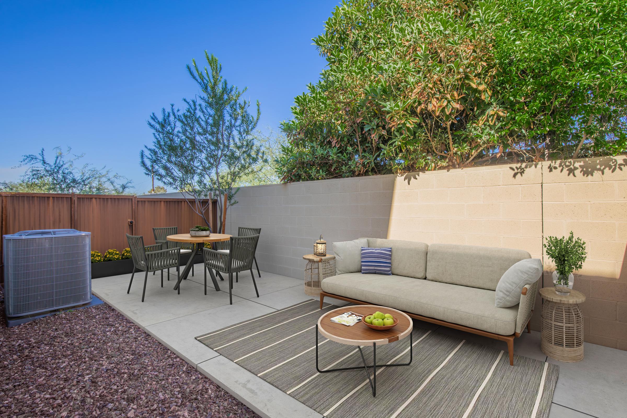 A cozy outdoor patio area featuring a light-colored sofa, a circular table with four black chairs, and a rug. The space is surrounded by greenery and a low wall, with an air conditioning unit in the corner, creating a serene atmosphere for relaxation or dining.