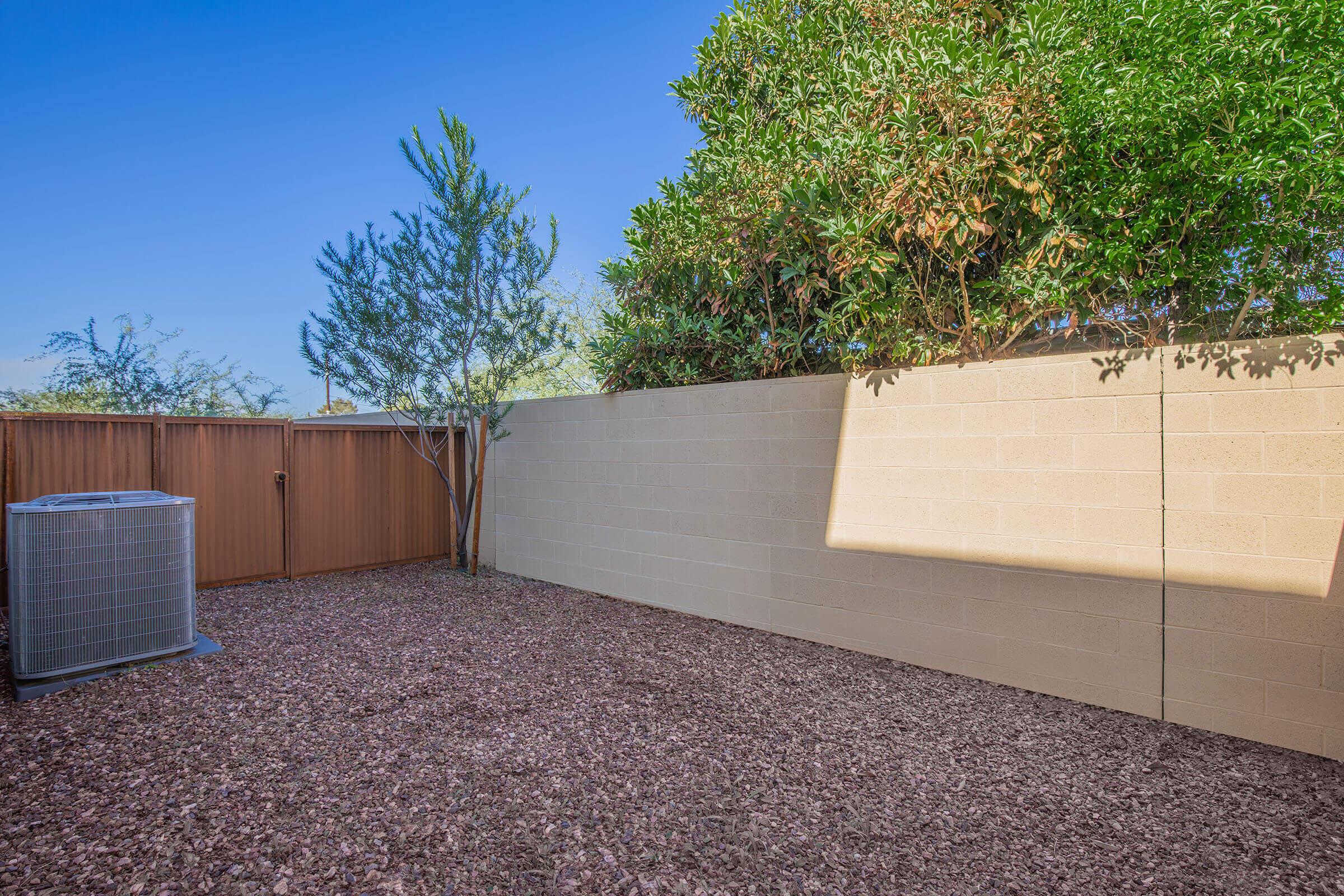 A small outdoor space featuring a gravel surface, a concrete block wall, and a hedge of greenery. An air conditioning unit is positioned in the corner, with clear blue skies above. The overall scene is tidy and designed for low maintenance.