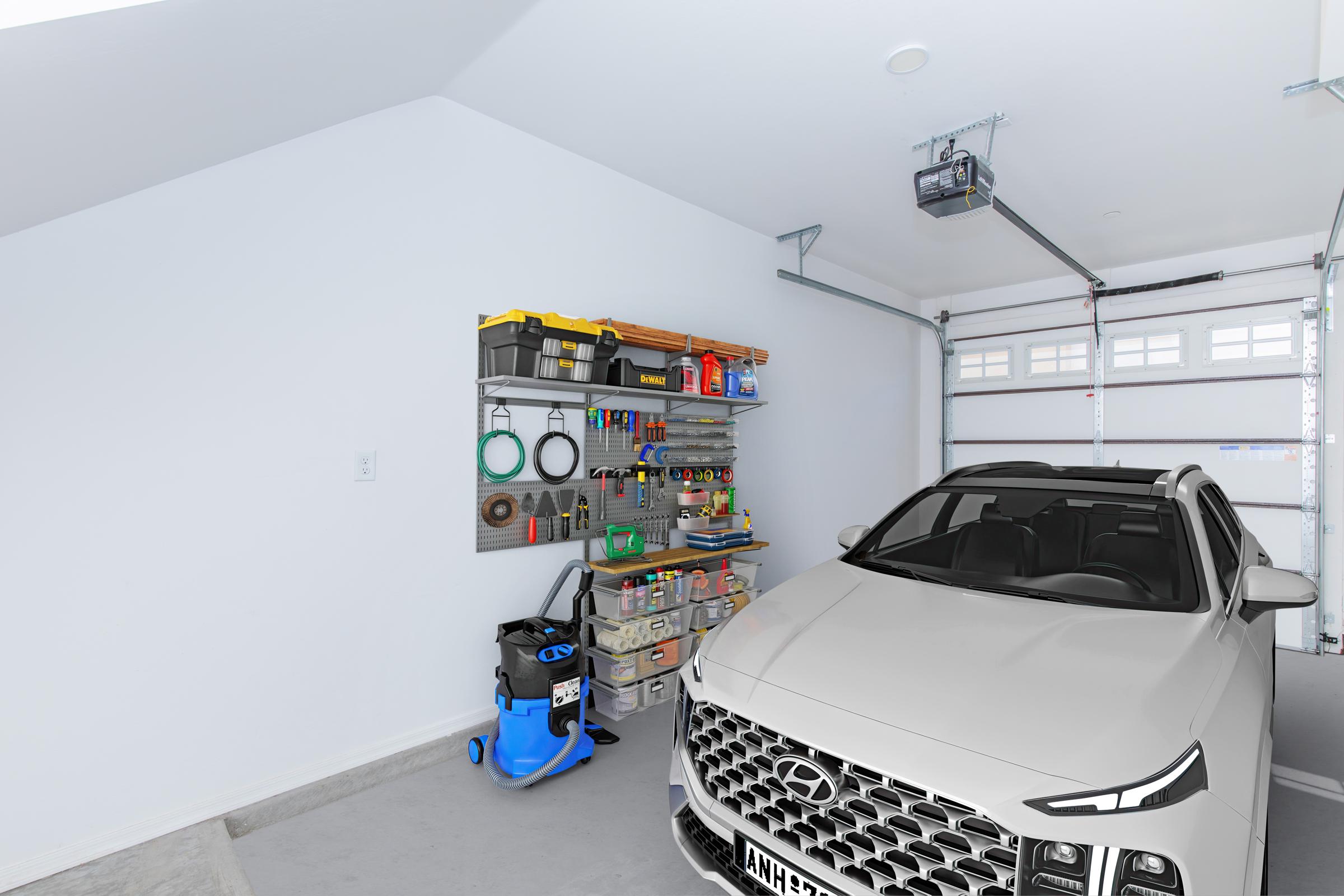A clean garage featuring a gray Hyundai SUV parked inside. The space includes a neatly organized storage wall with various tools, bins, and supplies. Additional items include a blue vacuum cleaner positioned on the floor. The garage door is partially open, allowing natural light to enter.