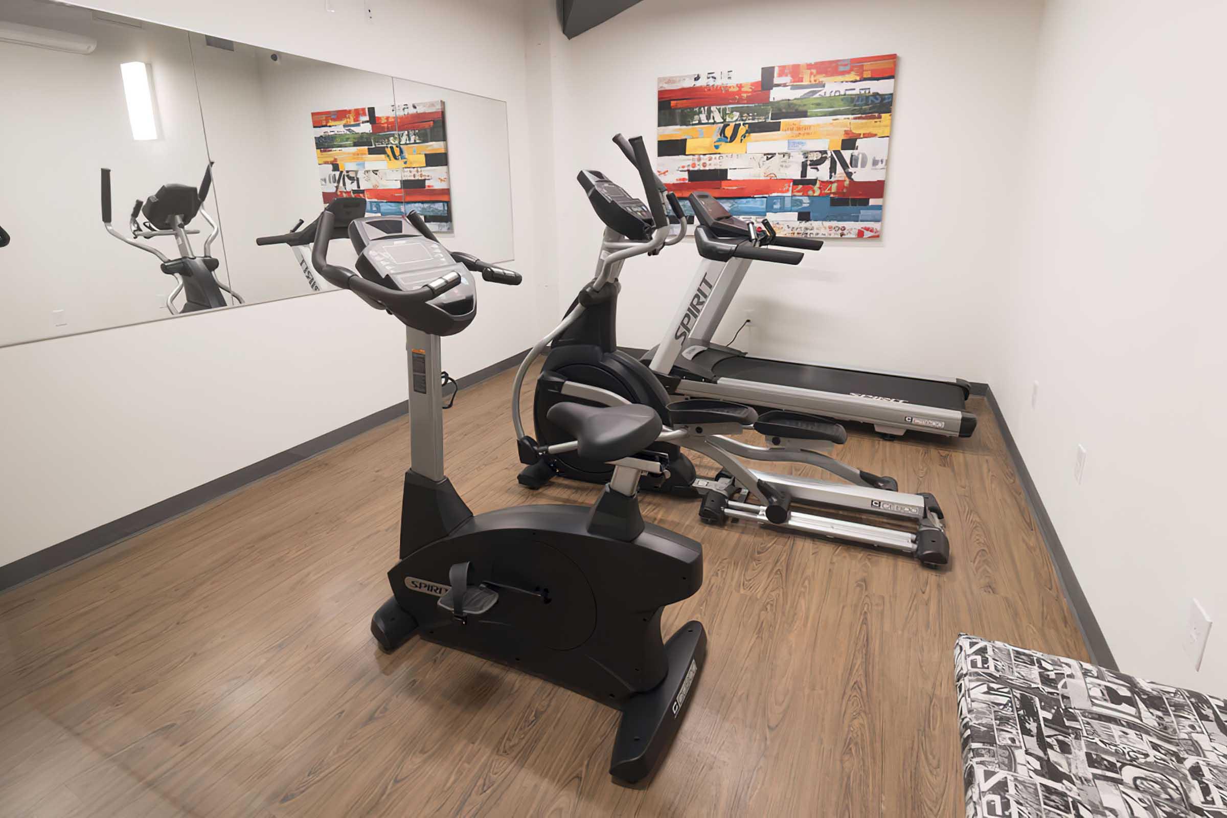 A small fitness room featuring exercise equipment, including an elliptical machine and a stationary bike. There is a large mirror on one wall and colorful artwork hanging on another. The flooring is wooden, and there is a patterned bench in the corner. The space is well-lit and organized for workouts.