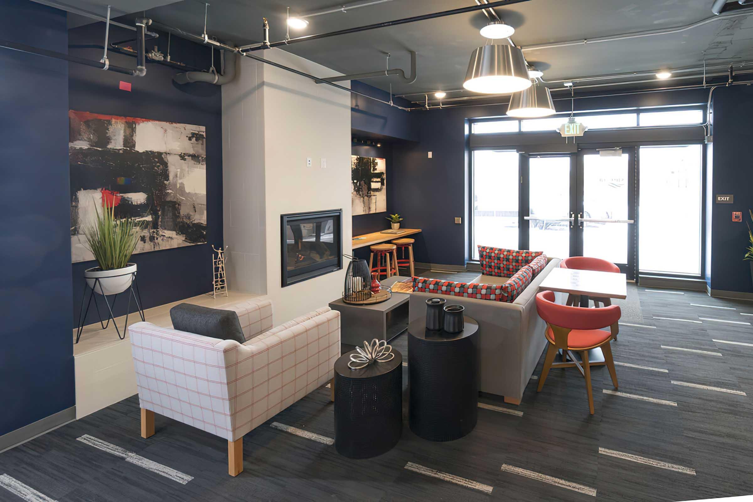 Modern lounge area featuring a mix of seating options including a gray sofa, a patterned chair, and a wooden table. A fireplace is set into the wall, and large windows provide natural light. Decorative plants and artwork enhance the contemporary decor. The floor is carpeted in gray and white patterns.