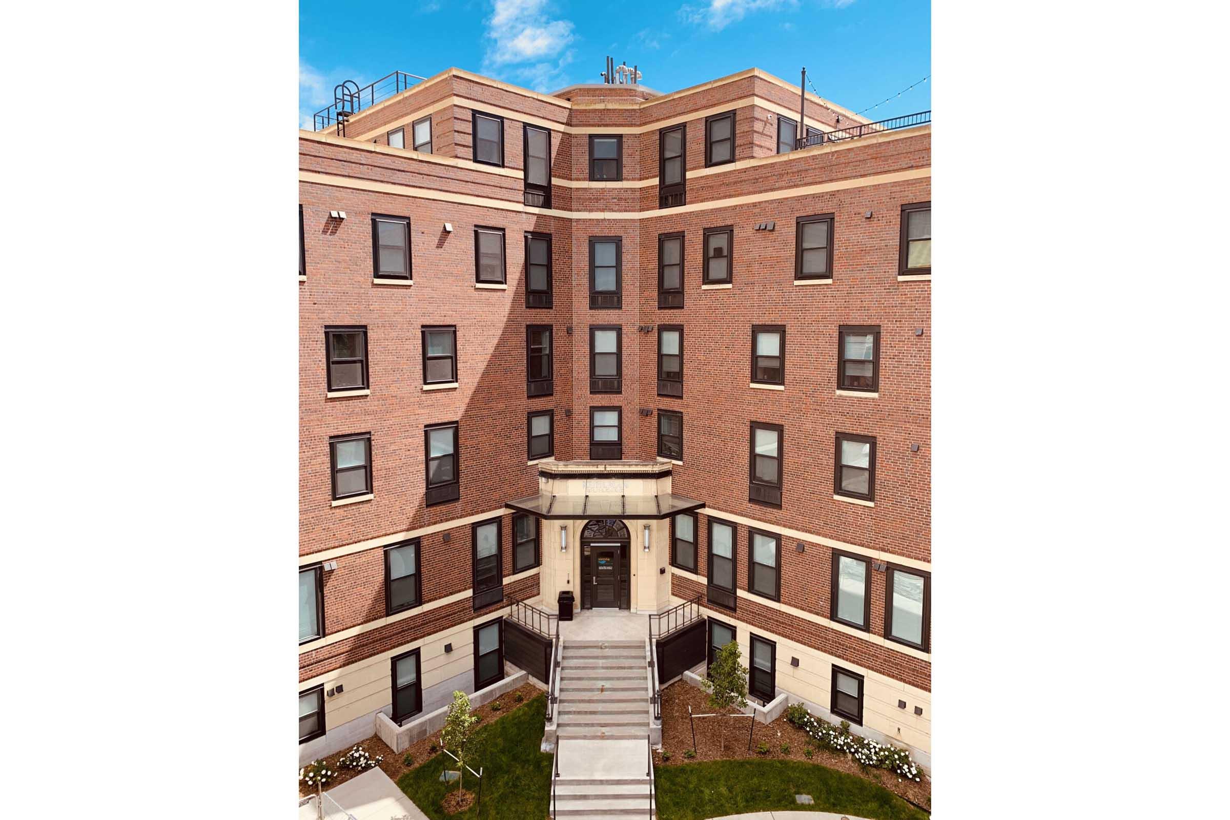 Exterior view of a large brick apartment building with multiple floors, featuring large windows and a central entrance with steps leading up to it. The building is surrounded by a landscaped area with grass and flowers, under a clear blue sky.