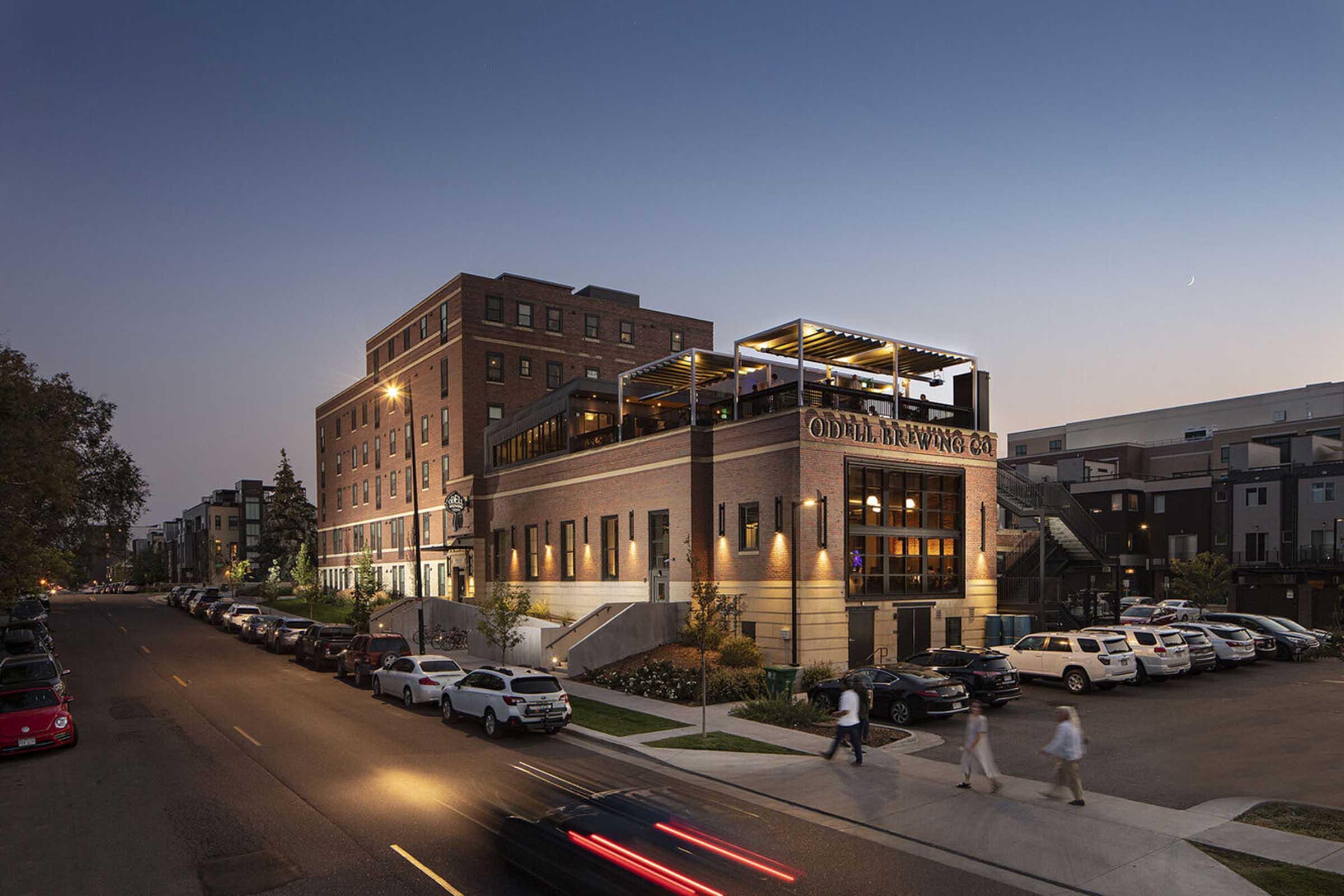 A modern brick building with a rooftop terrace, illuminated at dusk. Several cars are parked along the street, and a few people are walking nearby. The scene conveys a lively urban atmosphere, showcasing a blend of contemporary architecture and a welcoming environment.