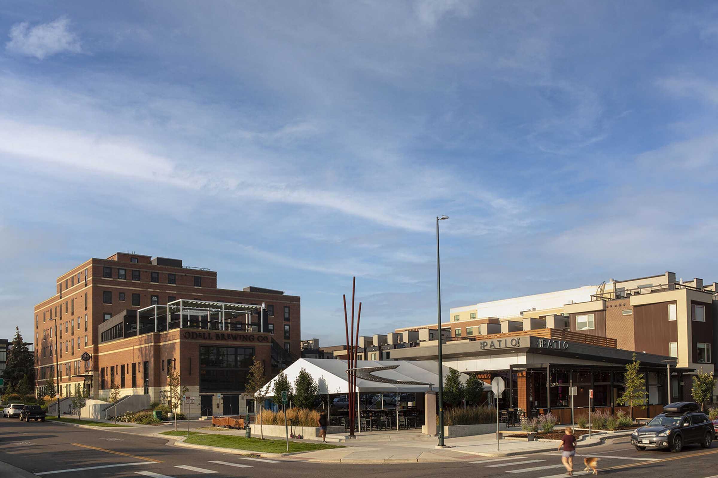 A street view featuring a modern building with a sign for "Patio" on the right and a larger, brick structure labeled "Canal Paving Co." on the left. The scene is set against a clear blue sky, with a landscaped area in front and a few pedestrians walking along the sidewalk.