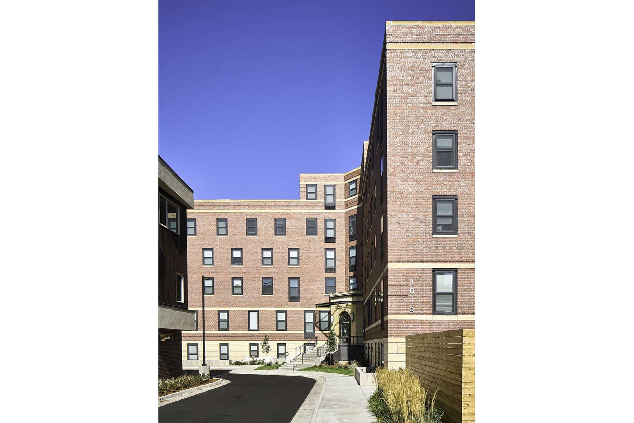 A modern multi-story brick building with multiple windows, featuring a side entrance and landscaped pathway. The sky above is clear and blue, and there are adjacent buildings with a contrasting design. A well-maintained area with greenery is visible in the foreground.